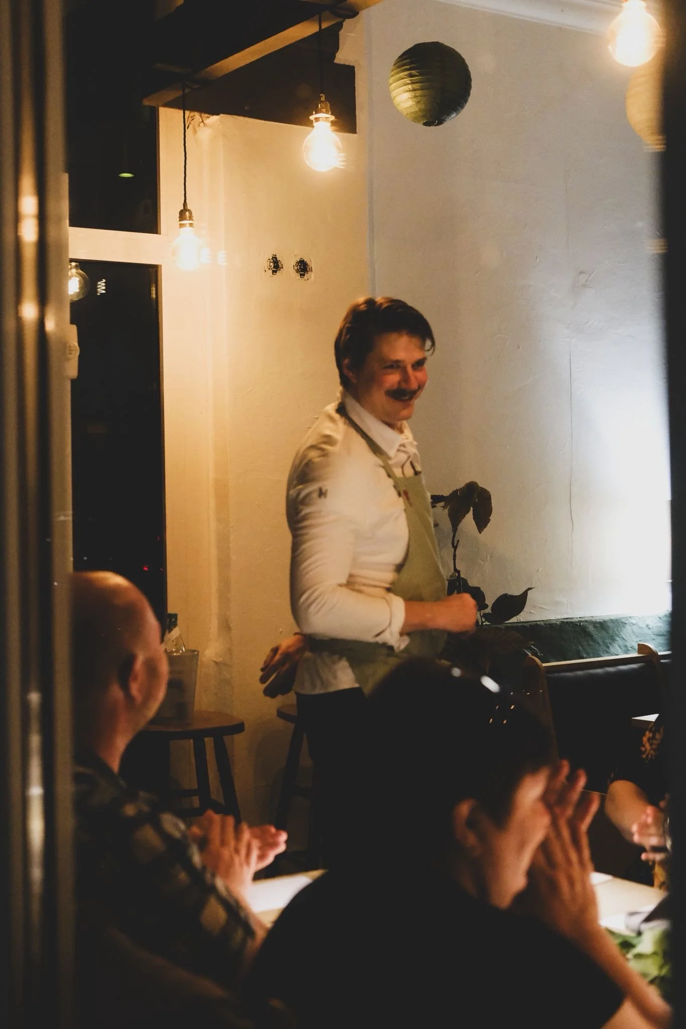 Ein lachender Koch erklärt den Gang, im Hintergrund sitzen mehrere Menschen in einem Restaurant, es gibt warmes Licht und Dekorationen, die Atmosphäre ist entspannt und freundlich.