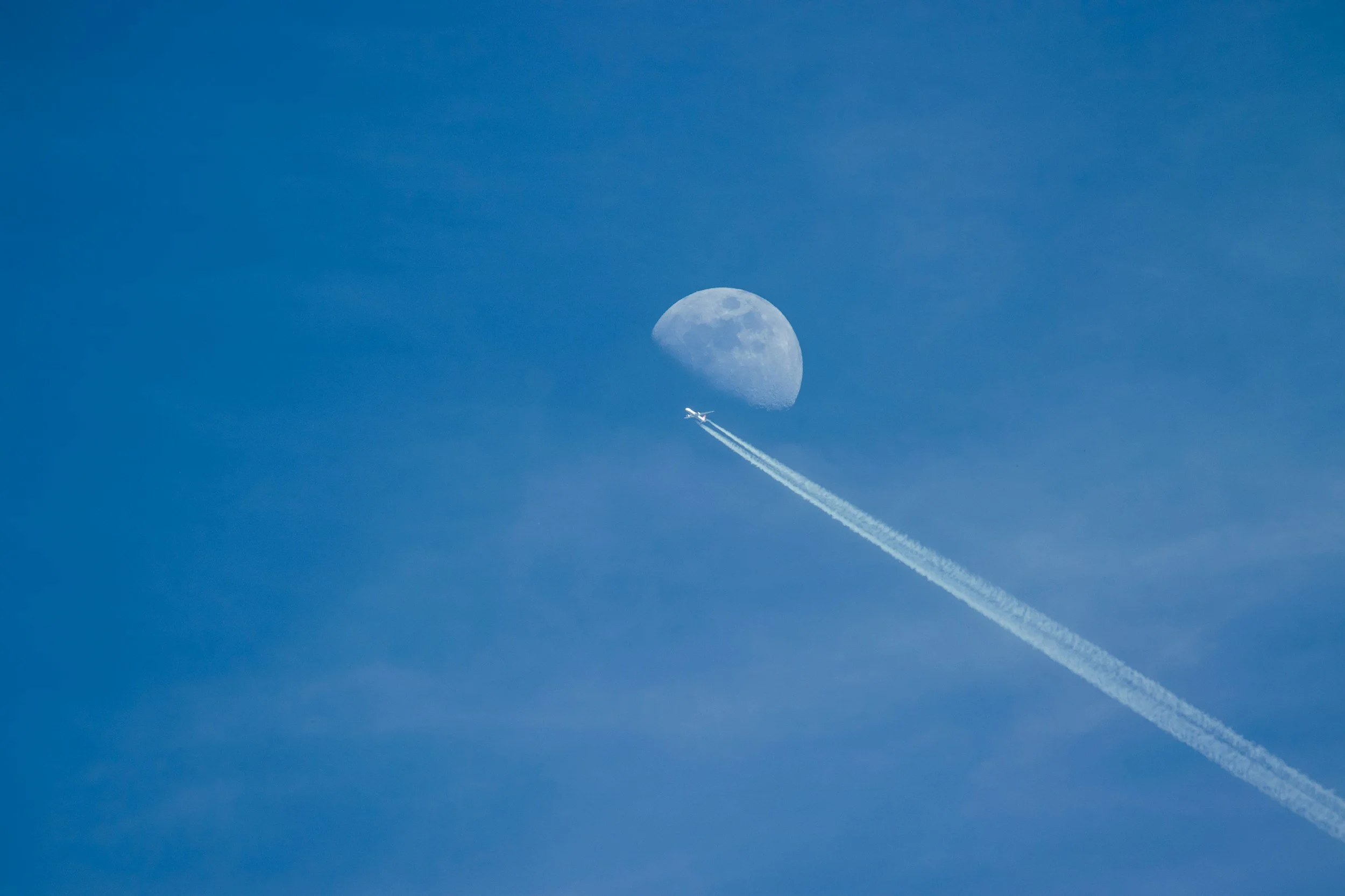An airplane flying in the sky leaving a contrail, with the moon visible in the background.