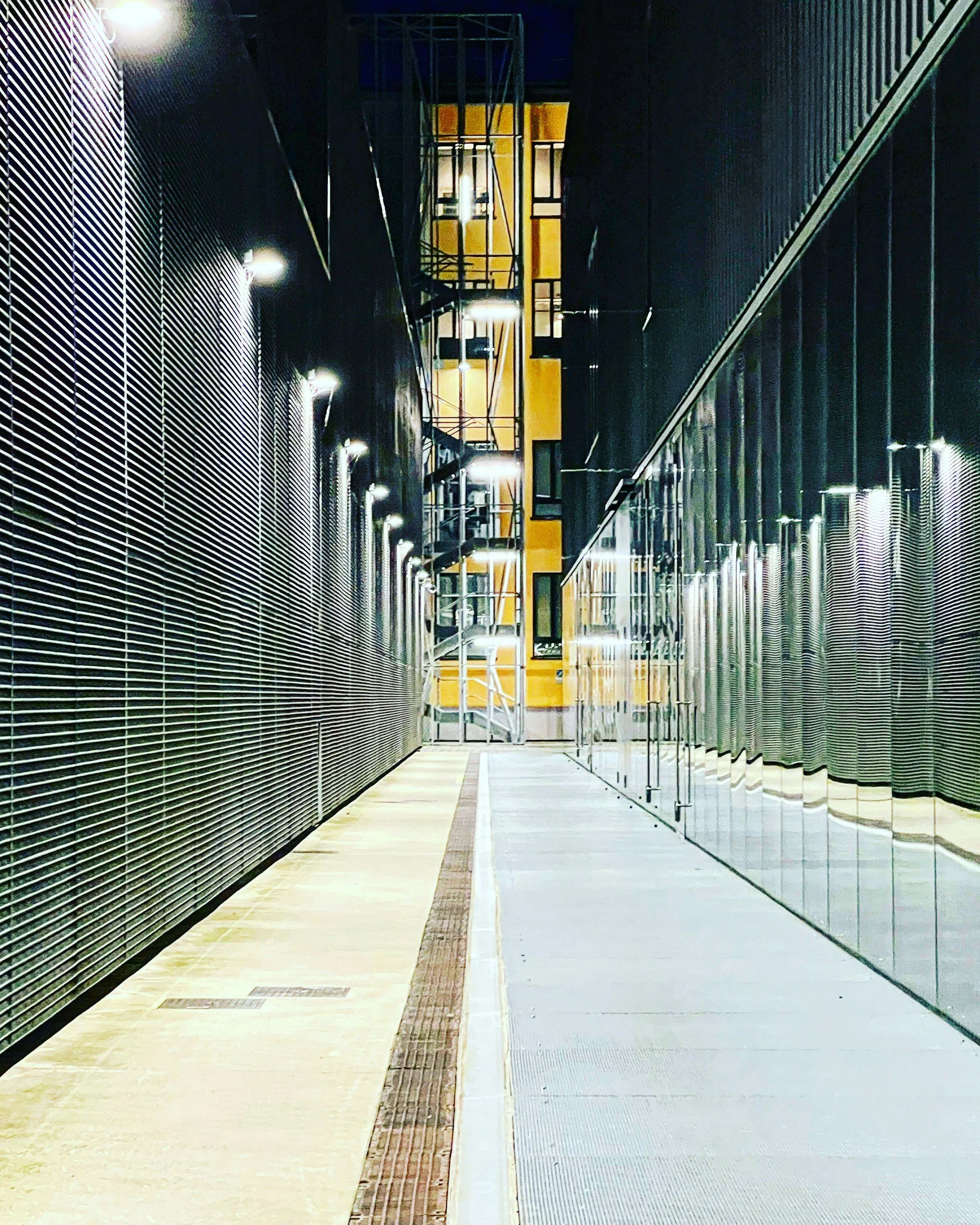 Empty corridor at night with modern metal architecture, illuminated by bright overhead lights. The corridor has metal side walls with glass on the right and a textured wall on the left. A yellow structure with stairs is visible at the end of the corridor.