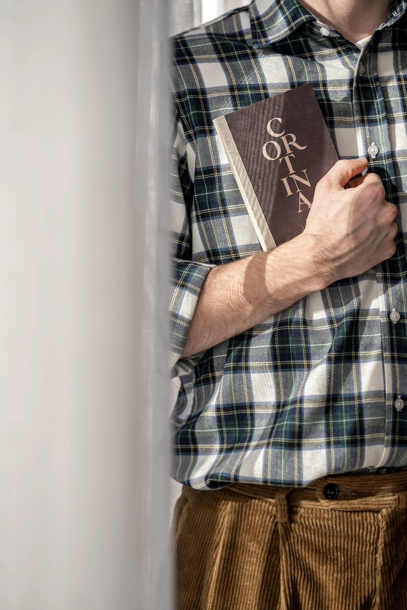 Persona che tiene un libro con la copertina ‘CORTINA’ e indossa una camicia a quadri sopra una maglietta bianca, pantaloni di velluto a coste marroni.