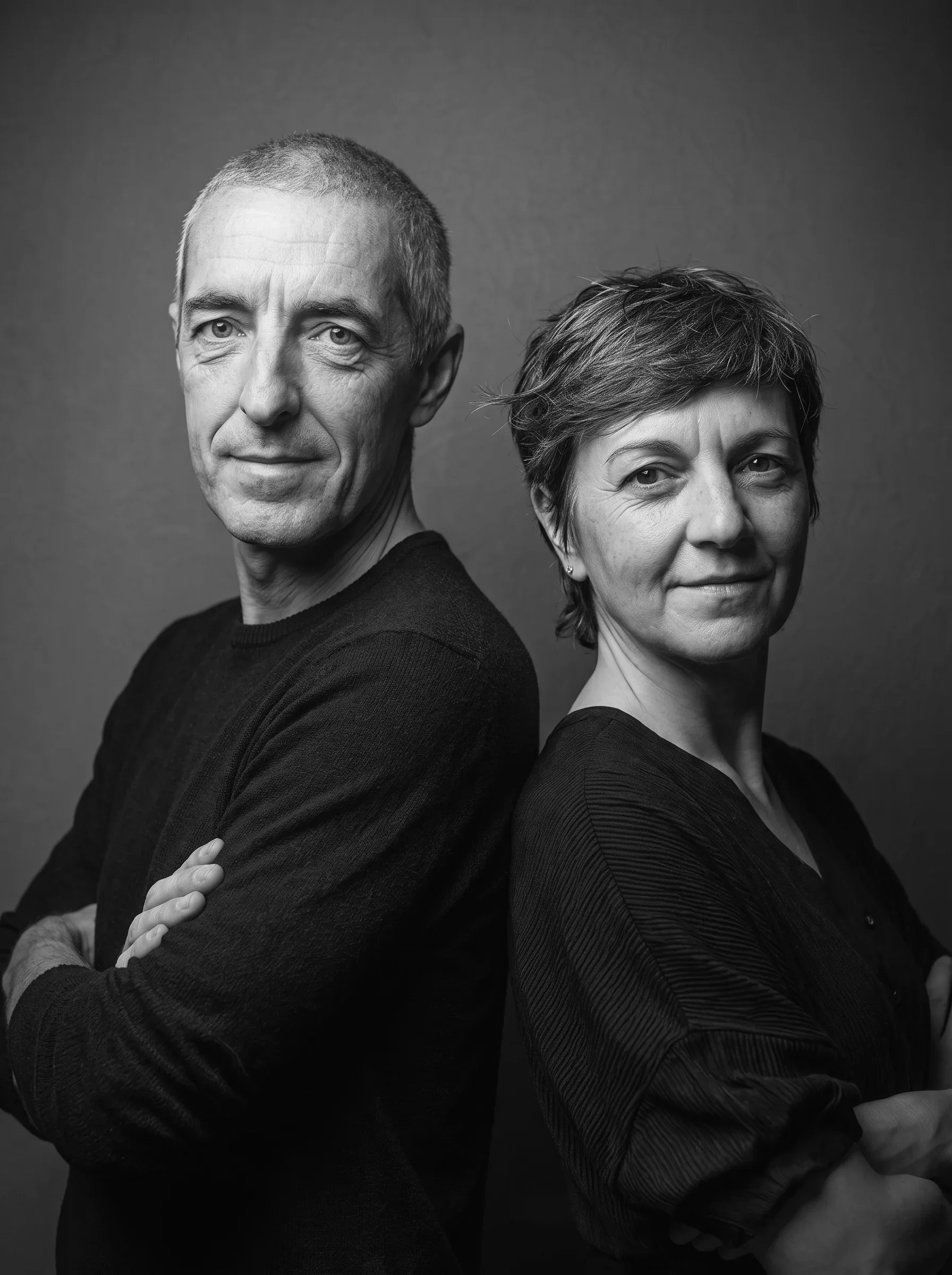 Una coppia di adulti, un uomo e una donna, si trova di fronte l'uno all'altro, con le braccia incrociate, in un'immagine in bianco e nero. L'uomo ha i capelli corti, grigi, e il volto leggermente agée, mentre la donna ha i capelli corti e mossi. Sono entrambi che sorridono leggermente, con sfondo neutro.