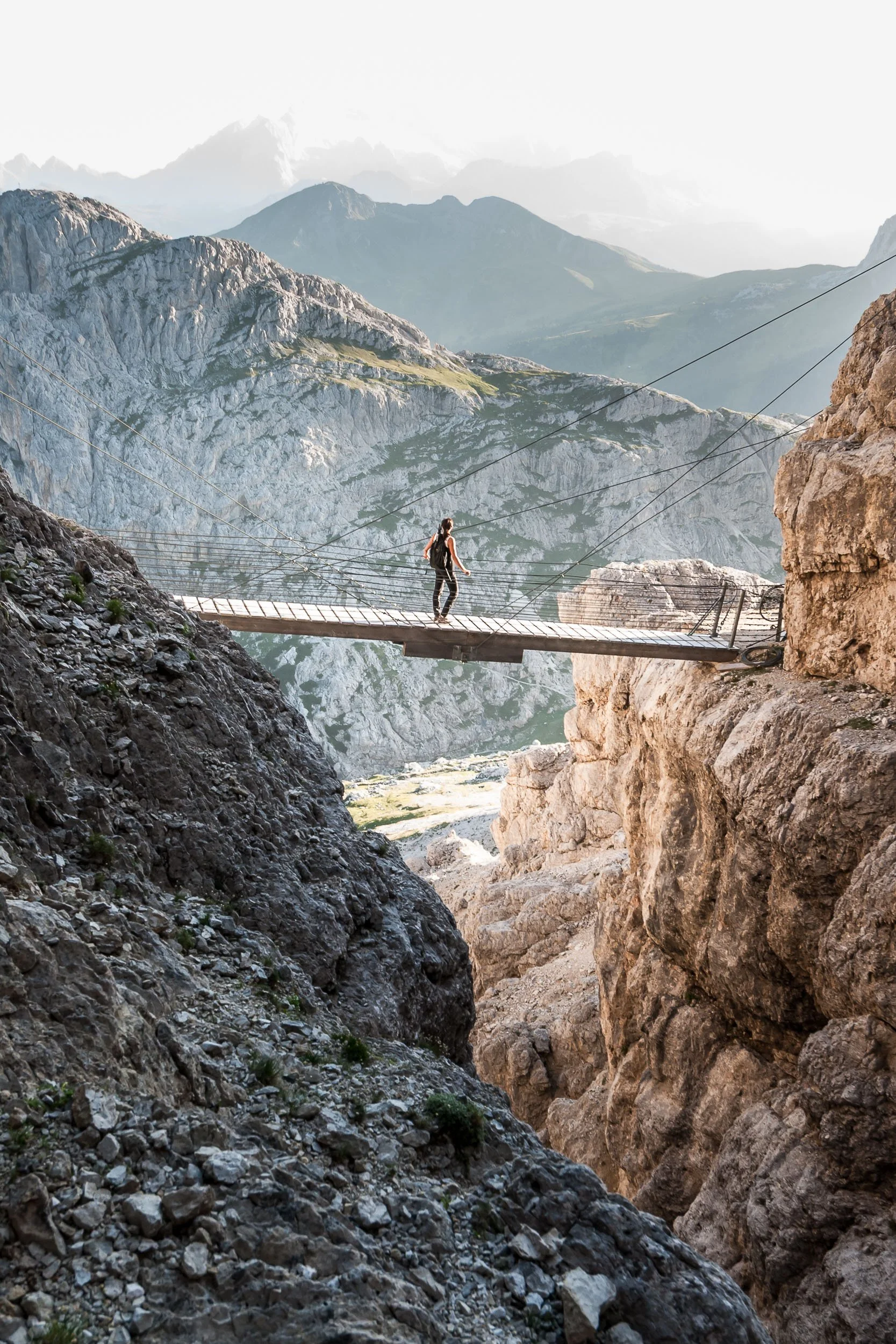 Un escursionista che attraversa un ponte di legno sospeso tra due pareti rocciose in un paesaggio montano con vette lontane e cielo sereno.
