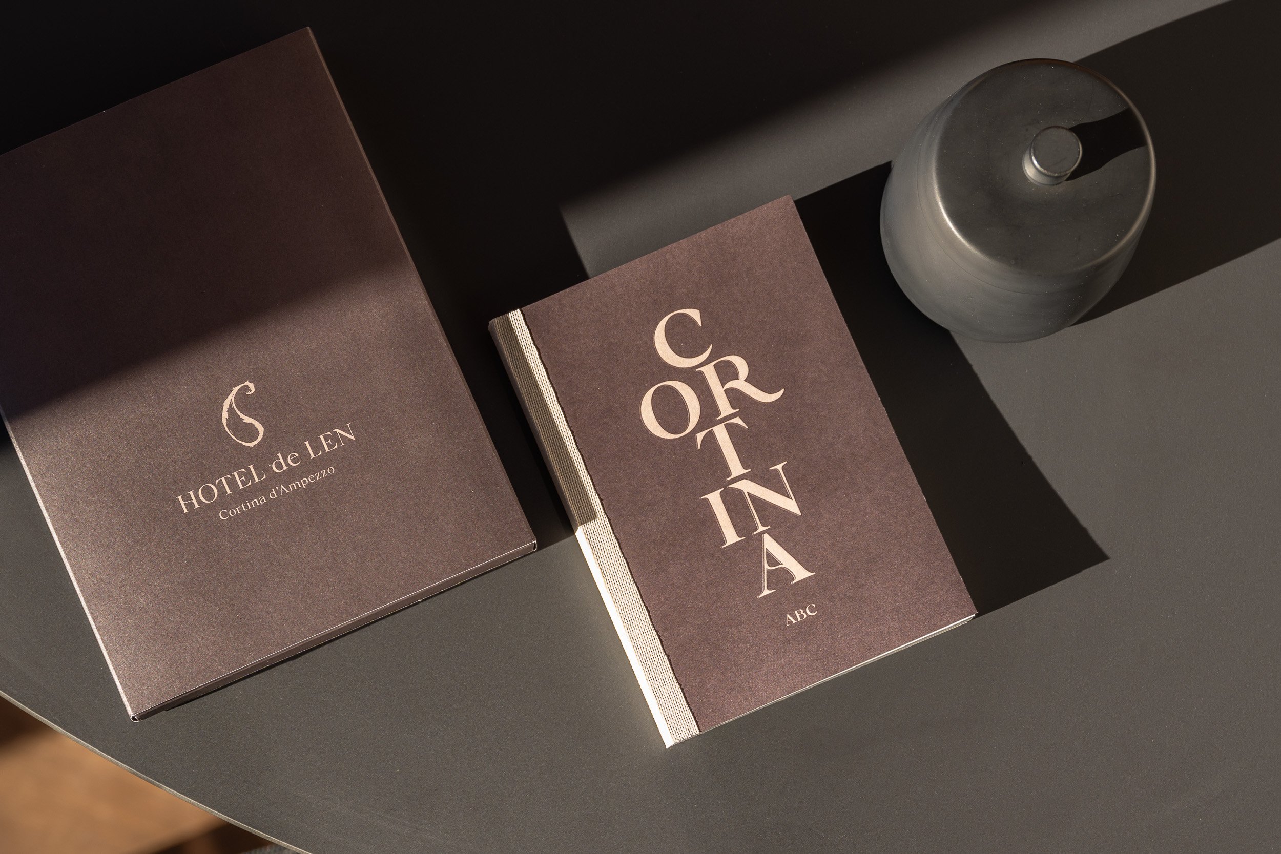 Libro marrone con titolo 'CORTINA' poggiato su una superficie nera accanto a un cofanetto marrone con scritto 'HOTEL de LEN Cortina d'Ampezzo' omaggio agli ospiti in accoglienza
