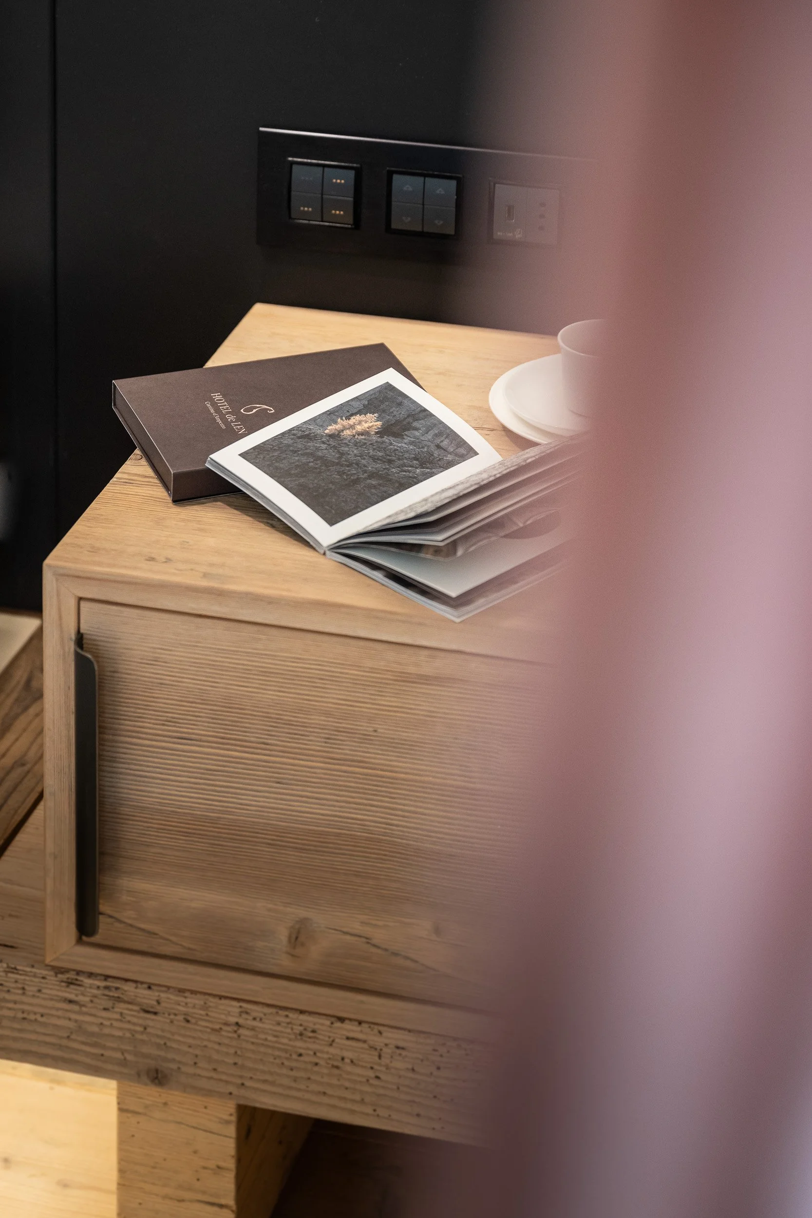 Tavolo di legno con un libro, una bevanda in una tazza e alcuni fogli o riviste. Sullo sfondo, una parete con prese e interruttori.