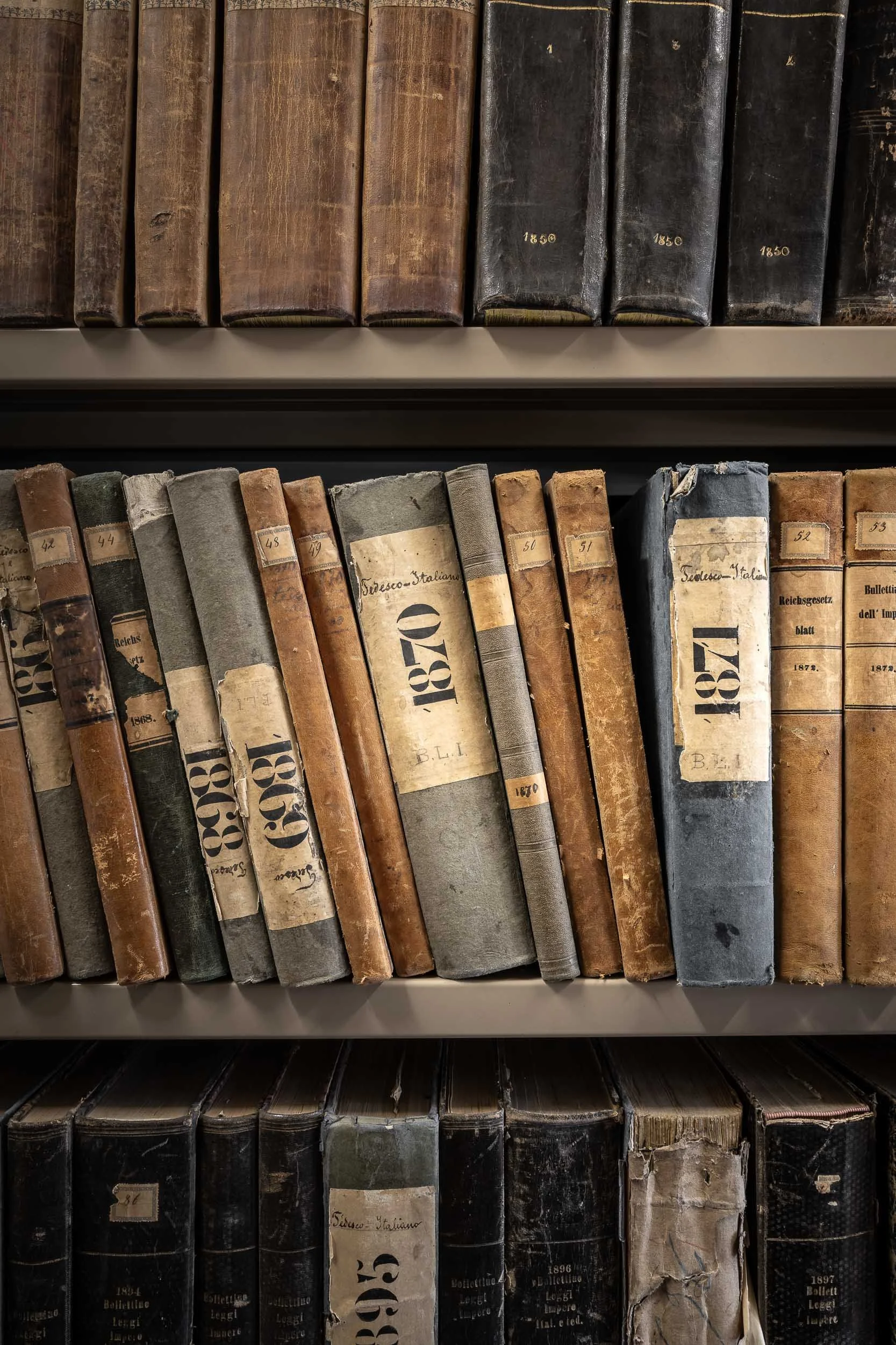 Libri antichi su scaffali, alcuni con copertine in pelle e etichette con numeri e date, patrimonio dell'Archivio del Comune di Cortina d'Ampezzo.