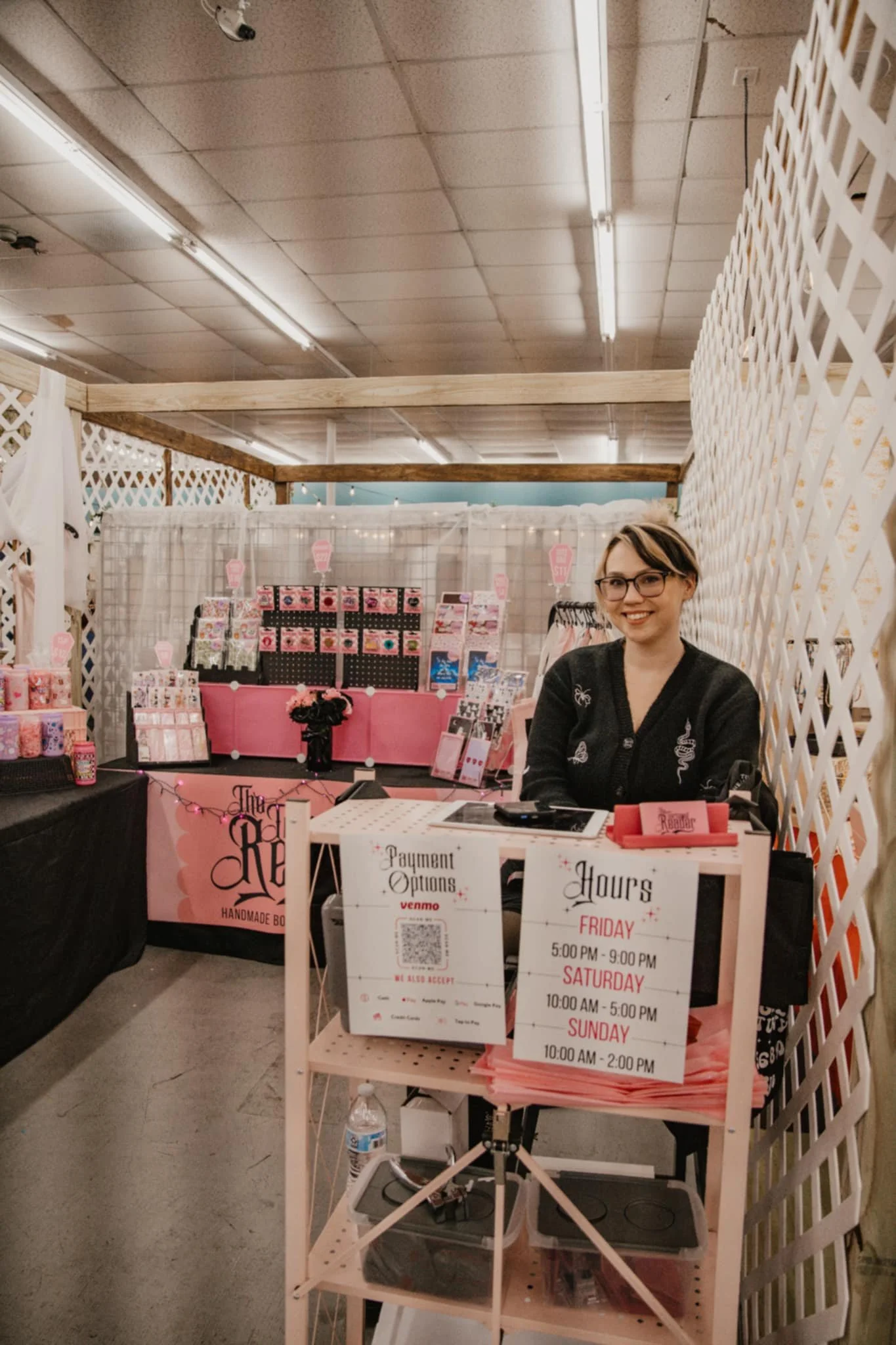 A woman with glasses and short hair sitting at a booth with a pink sign  an indoor market or craft fair. Behind her are displays of pink and patterned items, and a sign in front shows the hours of operation for a business, indicating it is open on Friday from 5:00 PM to 9:00 PM, Saturday from 10:00 AM to 5:00 PM, and Sunday from 10:00 AM to 2:00 PM.
