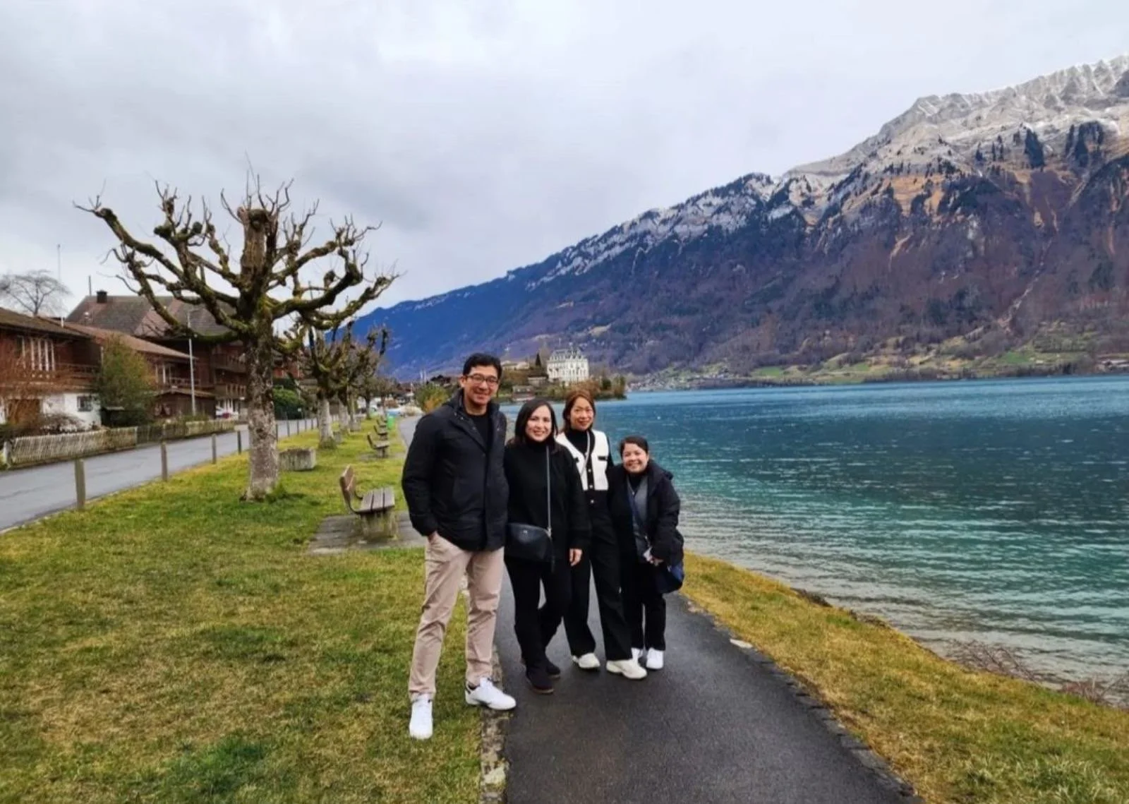 Four people standing on a paved path by a lake, with mountains in the background. There are leafless trees on the side of the path, and a row of houses along the lakeside. The sky is overcast.