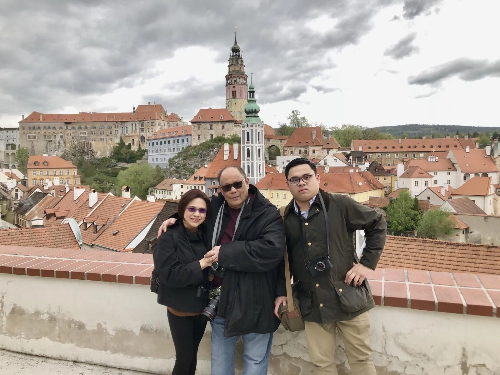 A family of three posing together outdoors with a European cityscape featuring red-tiled roofs, a tall tower, and a church steeple in the background, under a cloudy sky.