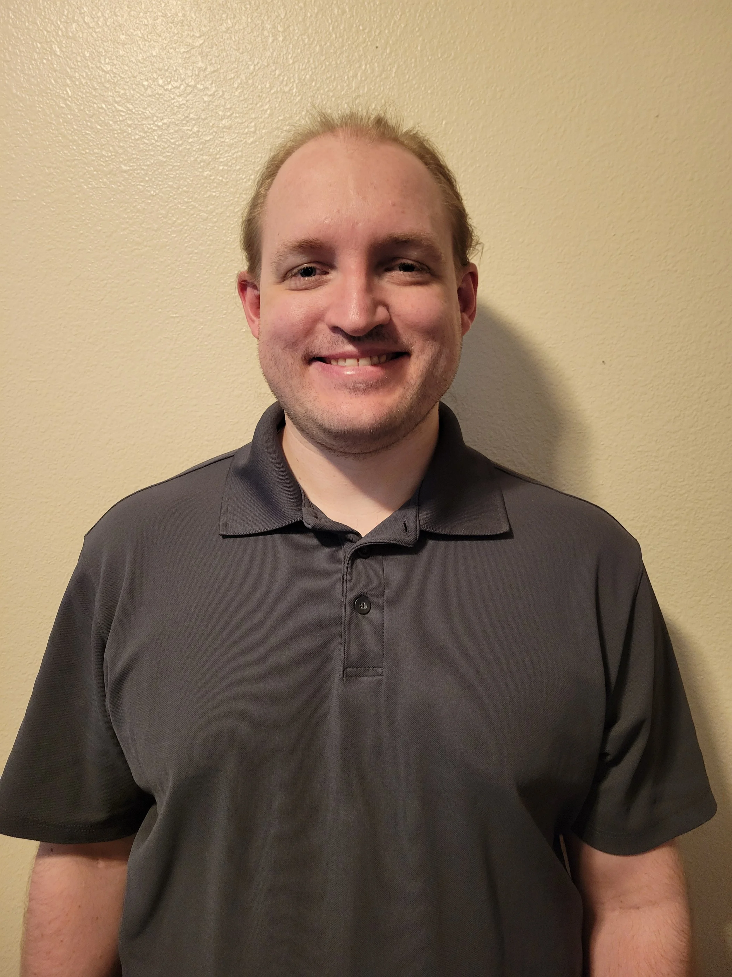 A man with light skin, light brown hair, and a slight beard wearing a dark gray polo shirt, smiling, standing against a beige wall.