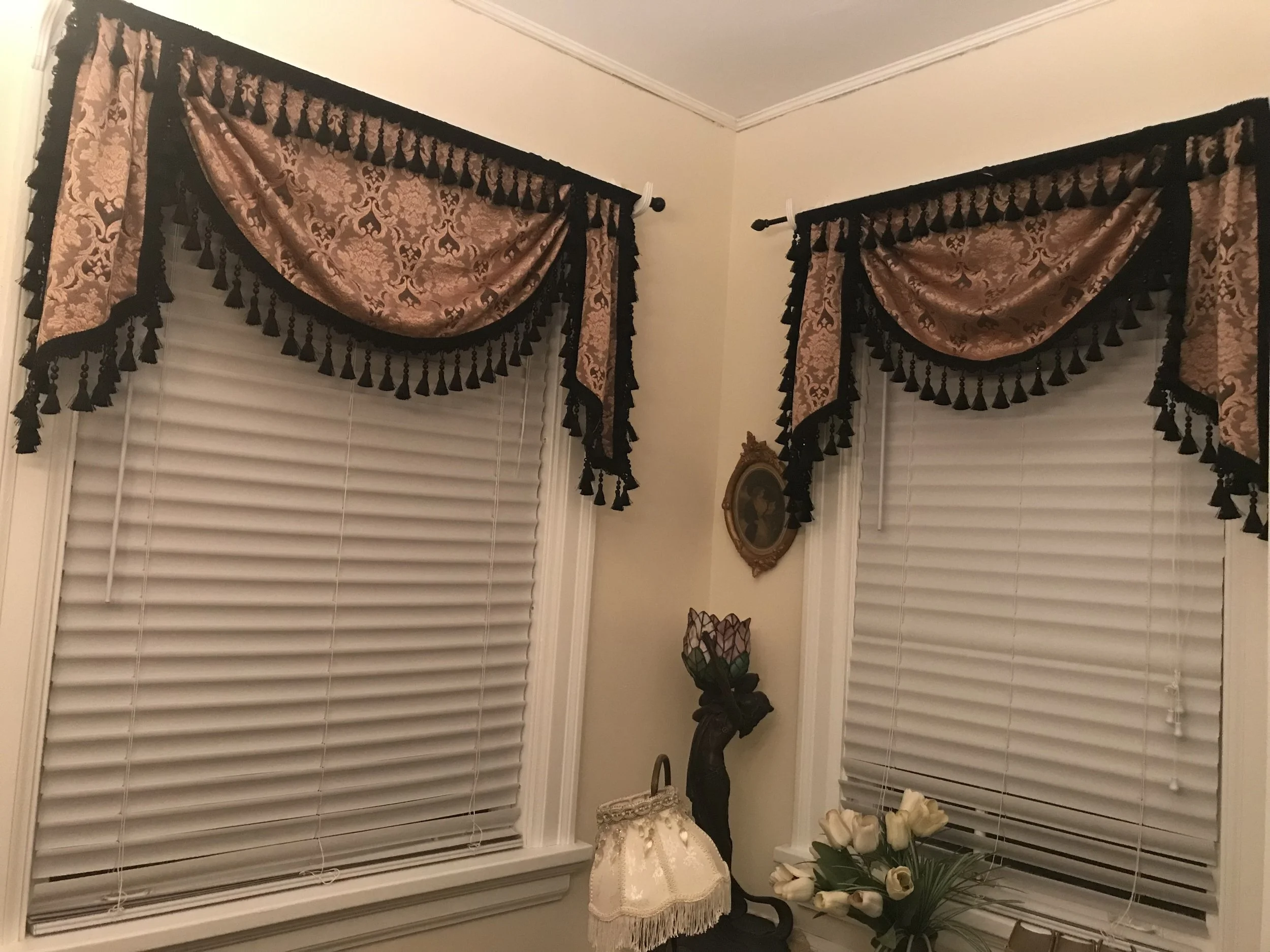 Decorative window curtains with floral patterns and black tassel trim, covering two windows with closed blinds, a framed portrait on the wall, a small table with a lace cloth, and a vase of white tulips.