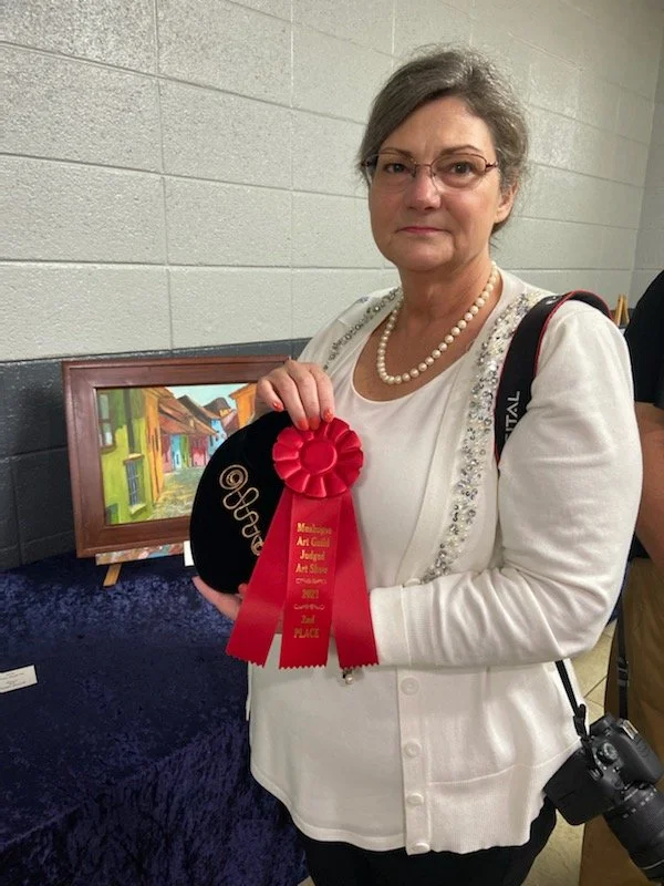 A woman holding a black award with a red ribbon that reads 'Second Place'. She is wearing glasses, a pearl necklace, and a white sweater with decorative trim. She stands next to a painting on a wooden easel in what appears to be an indoor art exhibit or competition.