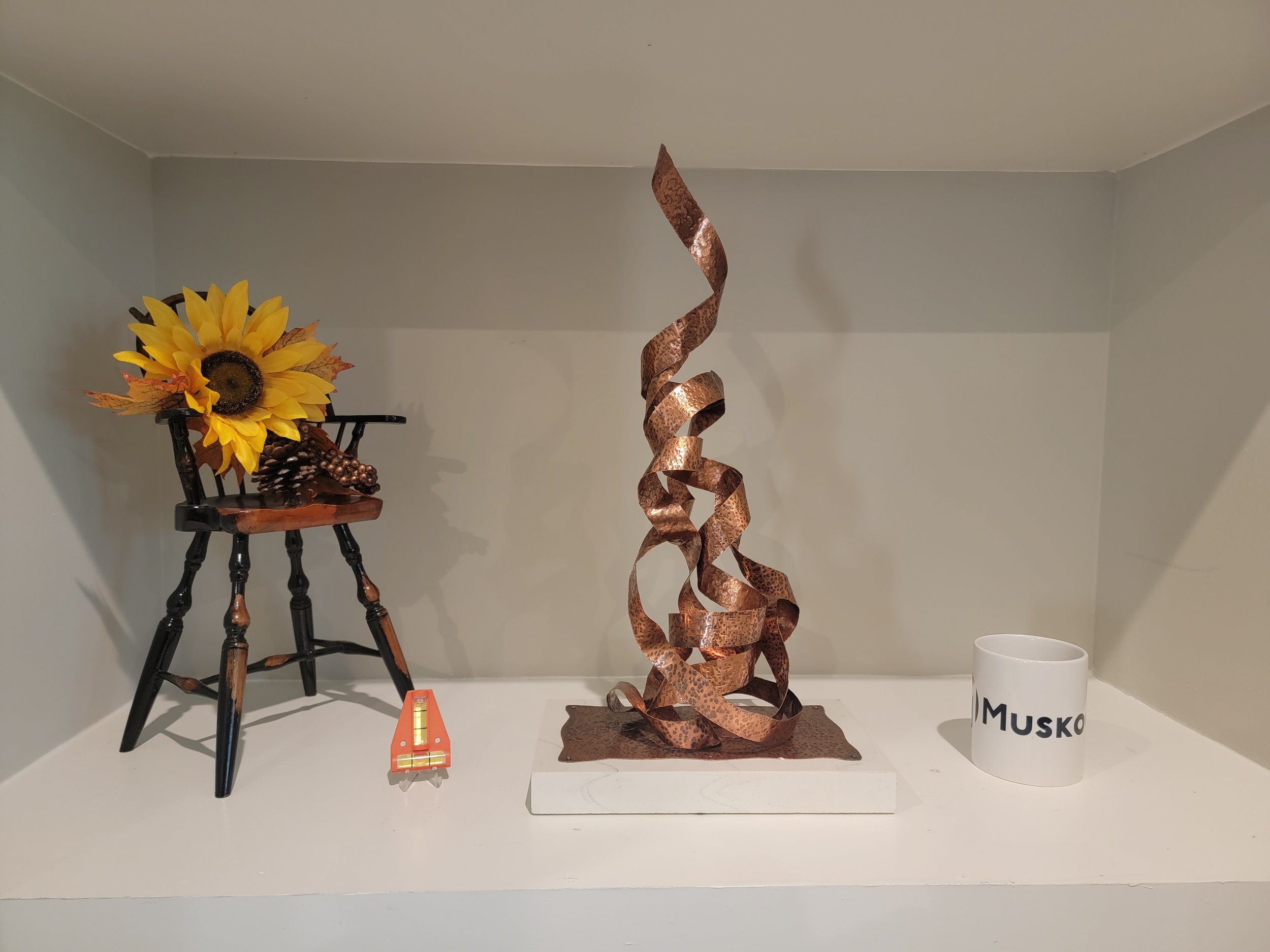 Decorative display on a white shelf with a sunflower accessory on a black stand, a twisted metal sculpture in copper color, a white mug with the name 'MUSK' partially visible, and a small orange box with a yellow level in front of the sunflower.