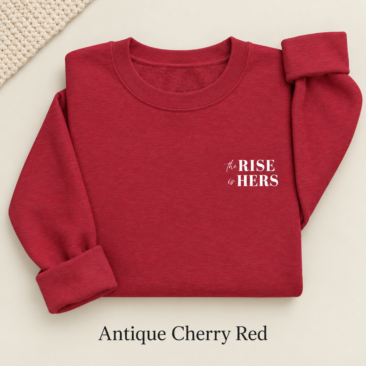 Antique Cherry Red The Rise Is Hers crewneck sweatshirt with an embroidered left-chest logo, folded on an ivory background.