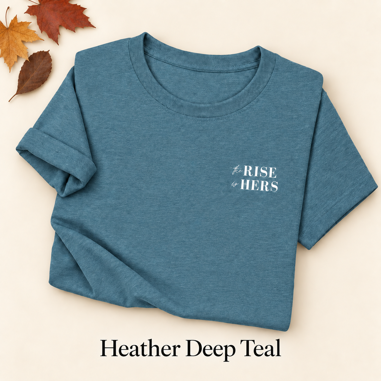 Heather Deep Teal The Rise Is Hers T-shirt with an embroidered left-chest logo, folded on an ivory background with autumn leaves.