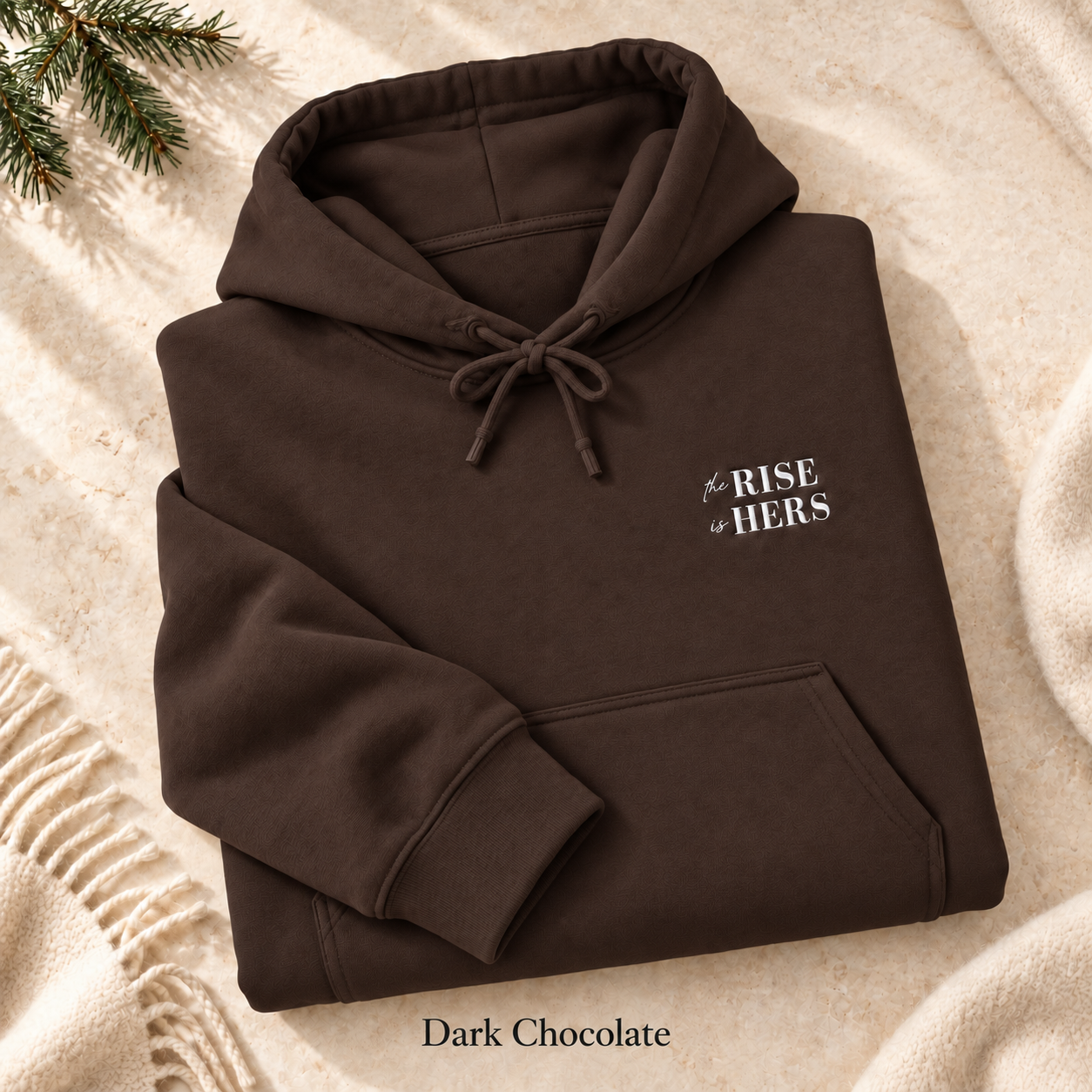 Dark Chocolate The Rise Is Hers hoodie with an embroidered left-chest logo, folded on cream fabric.