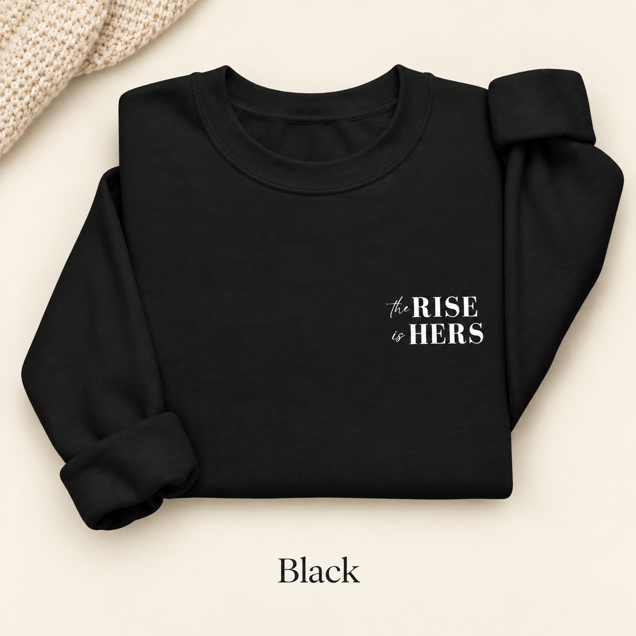 Black The Rise Is Hers crewneck sweatshirt with an embroidered left-chest logo, folded on an ivory background.