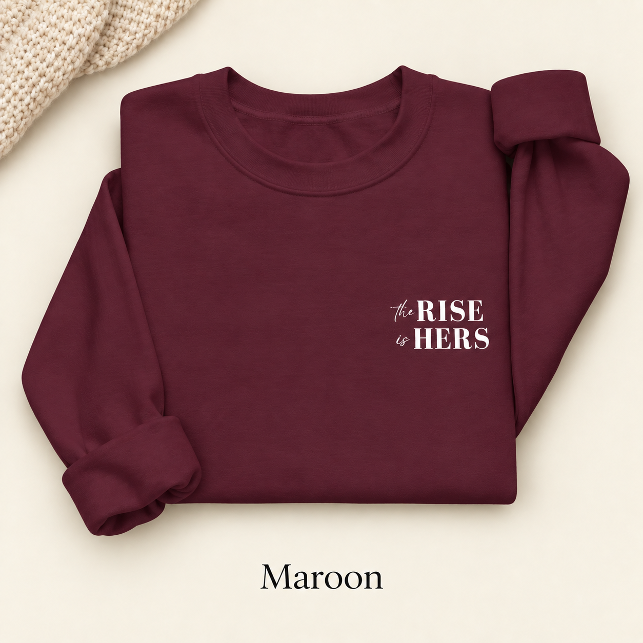 Maroon The Rise Is Hers crewneck sweatshirt with an embroidered left-chest logo, folded on an ivory background.