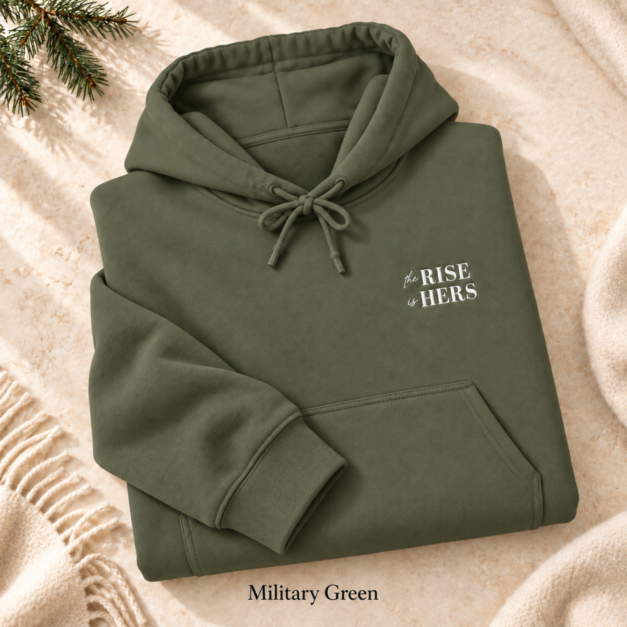 Military Green The Rise Is Hers hoodie with an embroidered left-chest logo, folded on cream fabric.