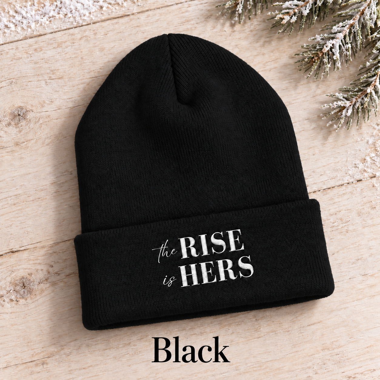 Black The Rise Is Hers cuffed knit beanie with an embroidered logo, displayed on a light background beside an evergreen branch.
