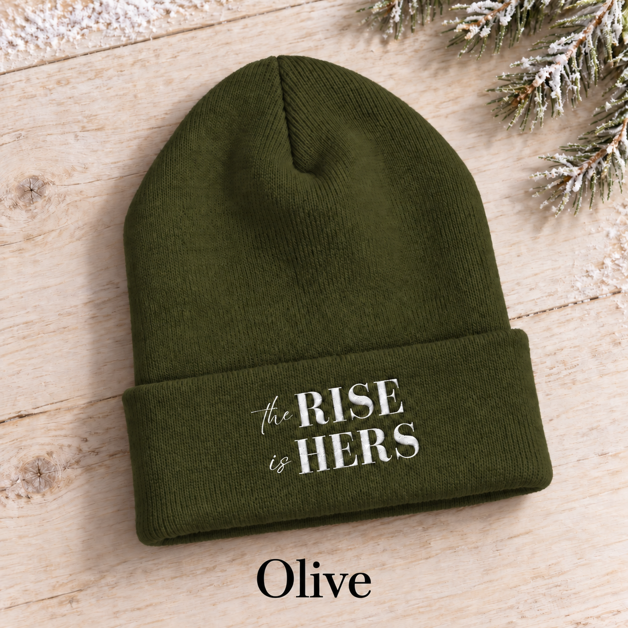Olive green The Rise Is Hers cuffed knit beanie with an embroidered logo, displayed on a light background beside an evergreen branch.