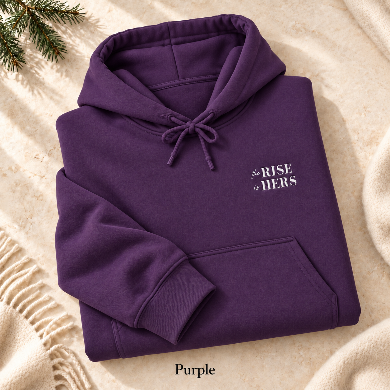 Purple The Rise Is Hers hoodie with an embroidered left-chest logo, folded on cream fabric.