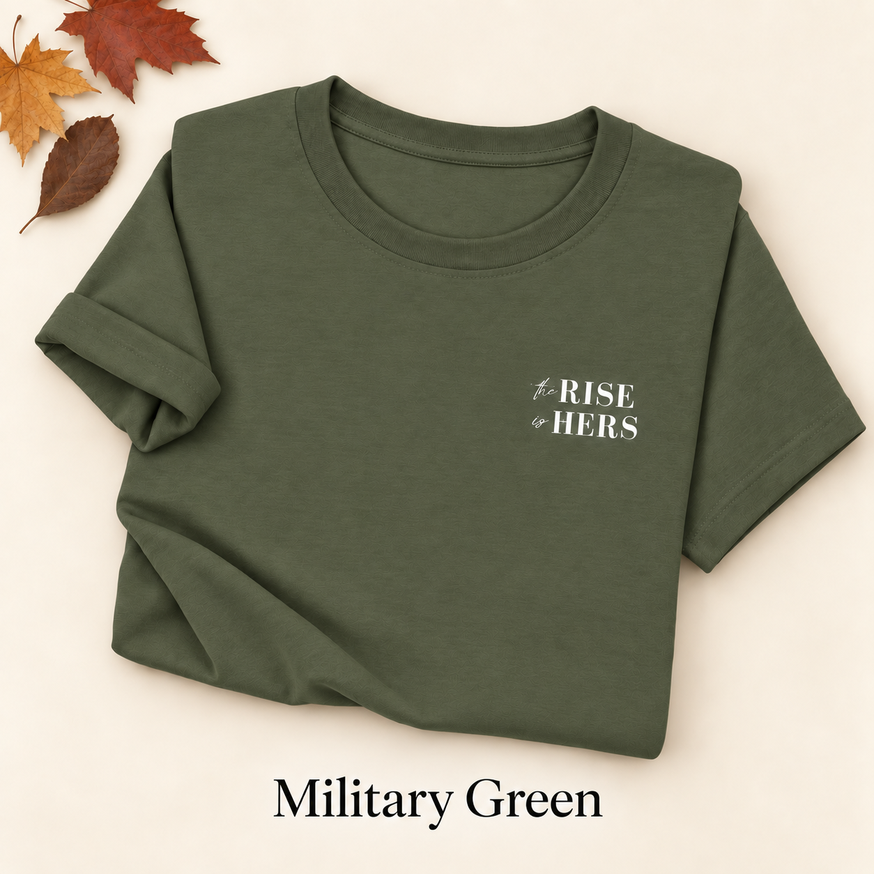 Military Green The Rise Is Hers T-shirt with an embroidered left-chest logo, folded on an ivory background with autumn leaves.
