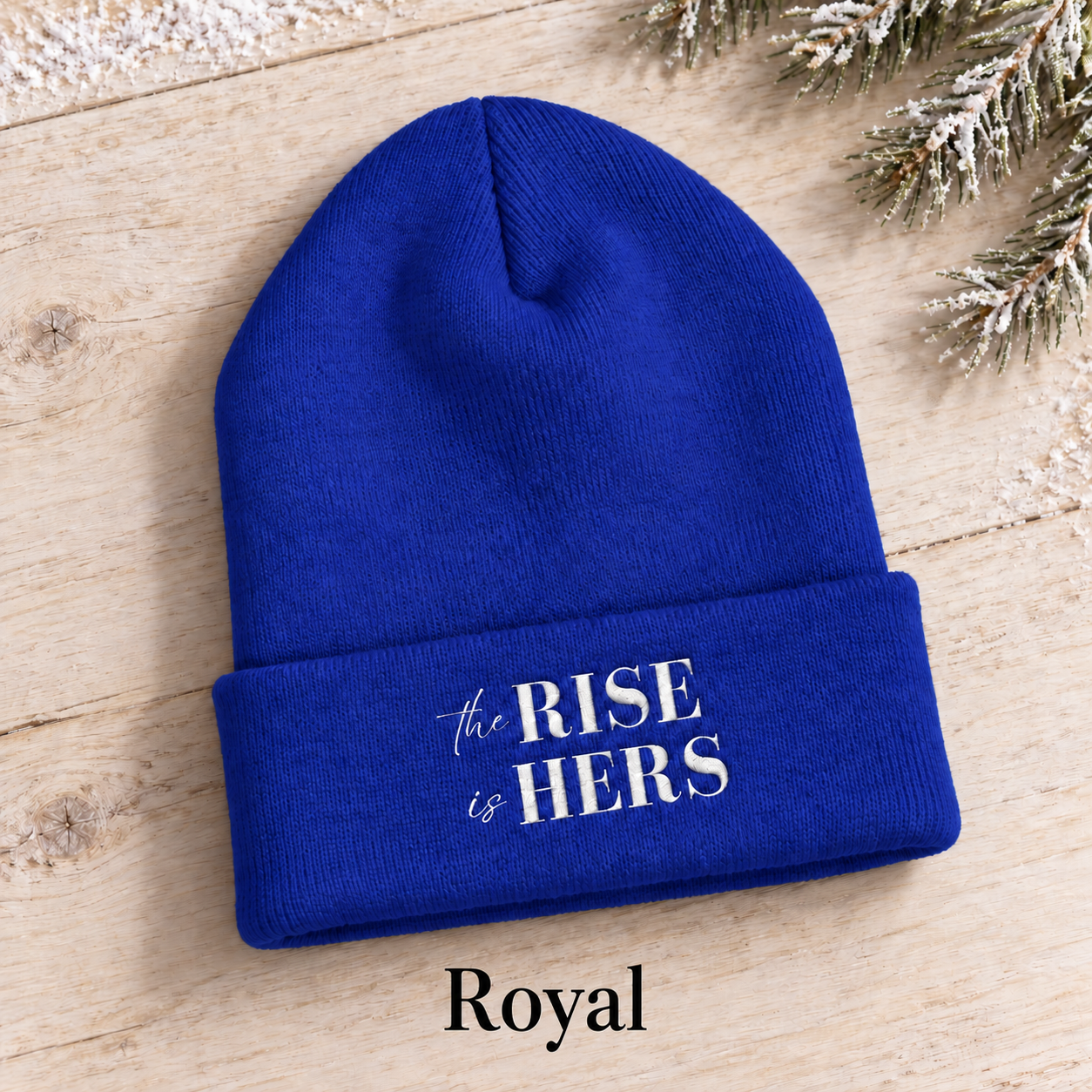 Royal blue The Rise Is Hers cuffed knit beanie with an embroidered logo, displayed on a light background beside an evergreen branch.