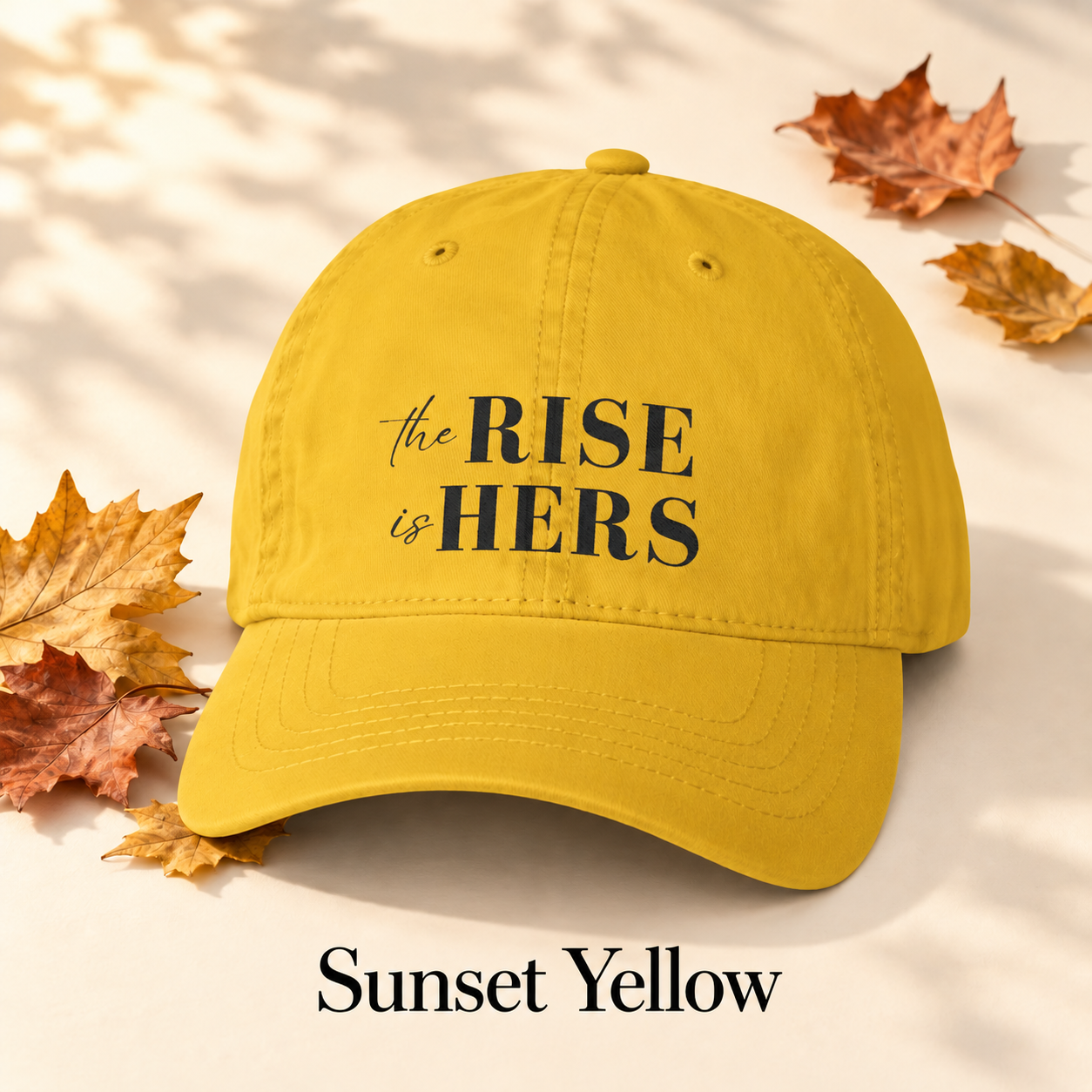 Sunset Yellow The Rise Is Hers baseball hat with an embroidered front logo, displayed beside autumn leaves.