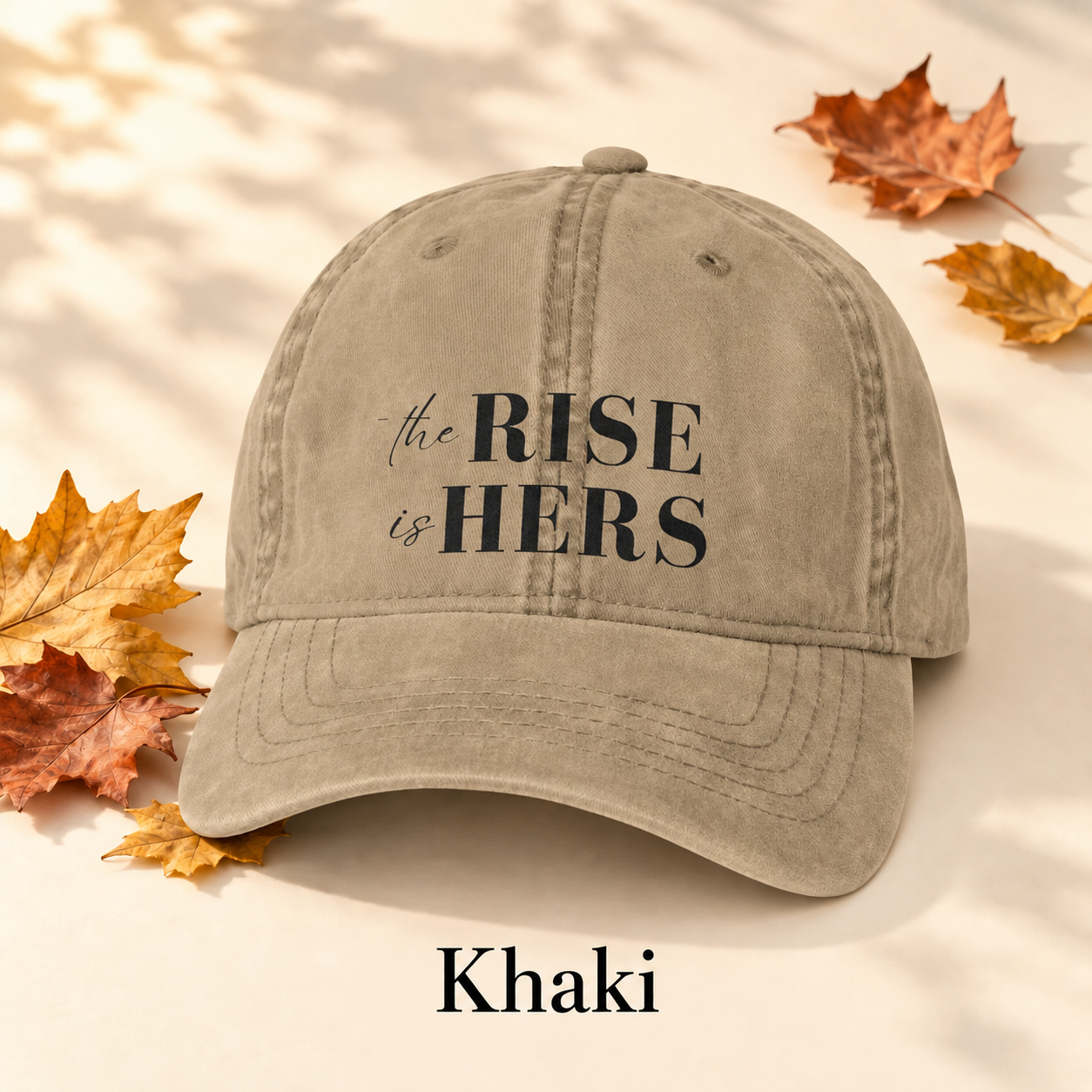 Khaki The Rise Is Hers baseball hat with an embroidered front logo, displayed beside autumn leaves.