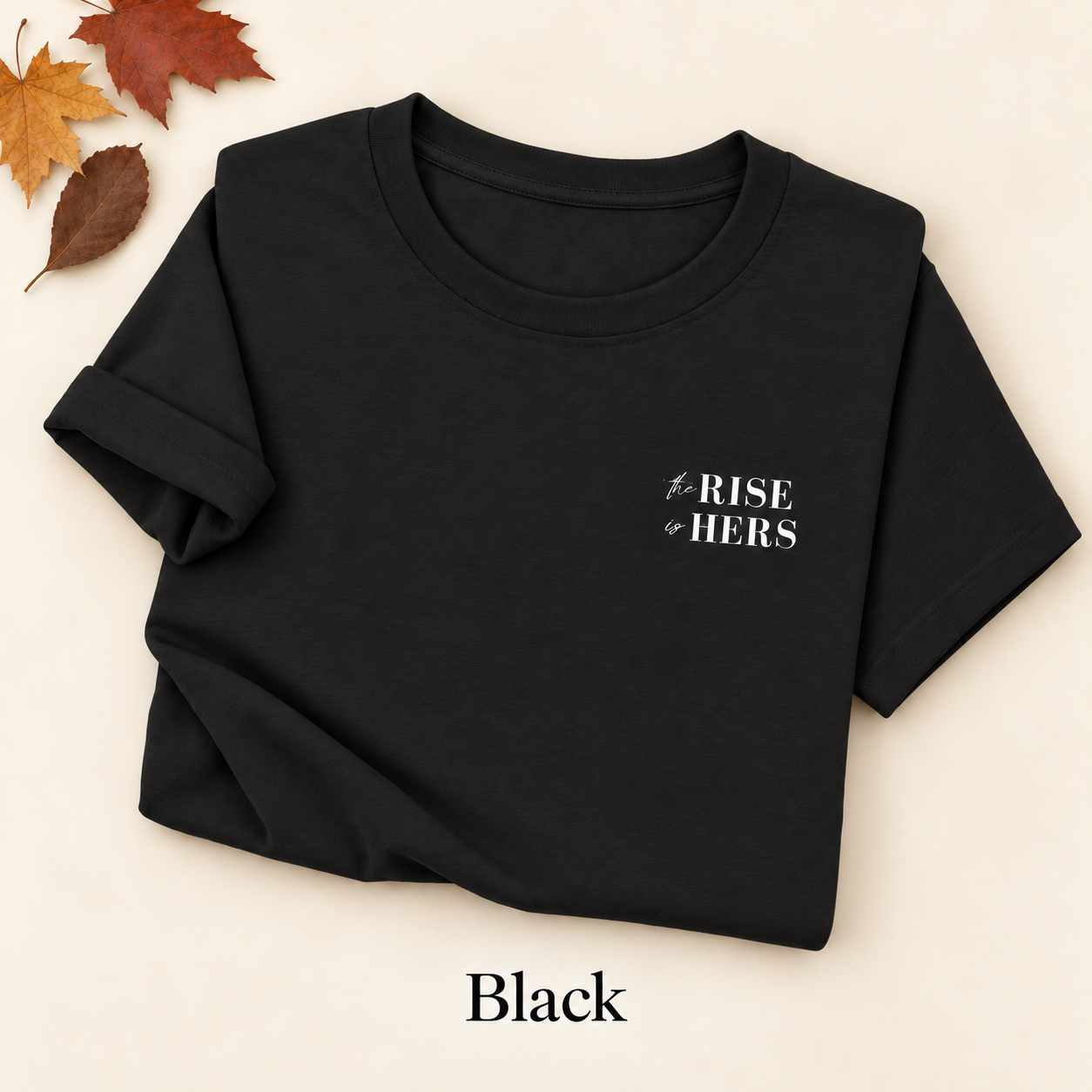 Black The Rise Is Hers T-shirt with an embroidered left-chest logo, folded on an ivory background with autumn leaves.