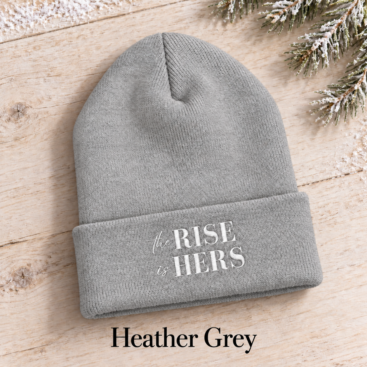 Heather Grey The Rise Is Hers cuffed knit beanie with an embroidered logo, displayed on a light background beside an evergreen branch.