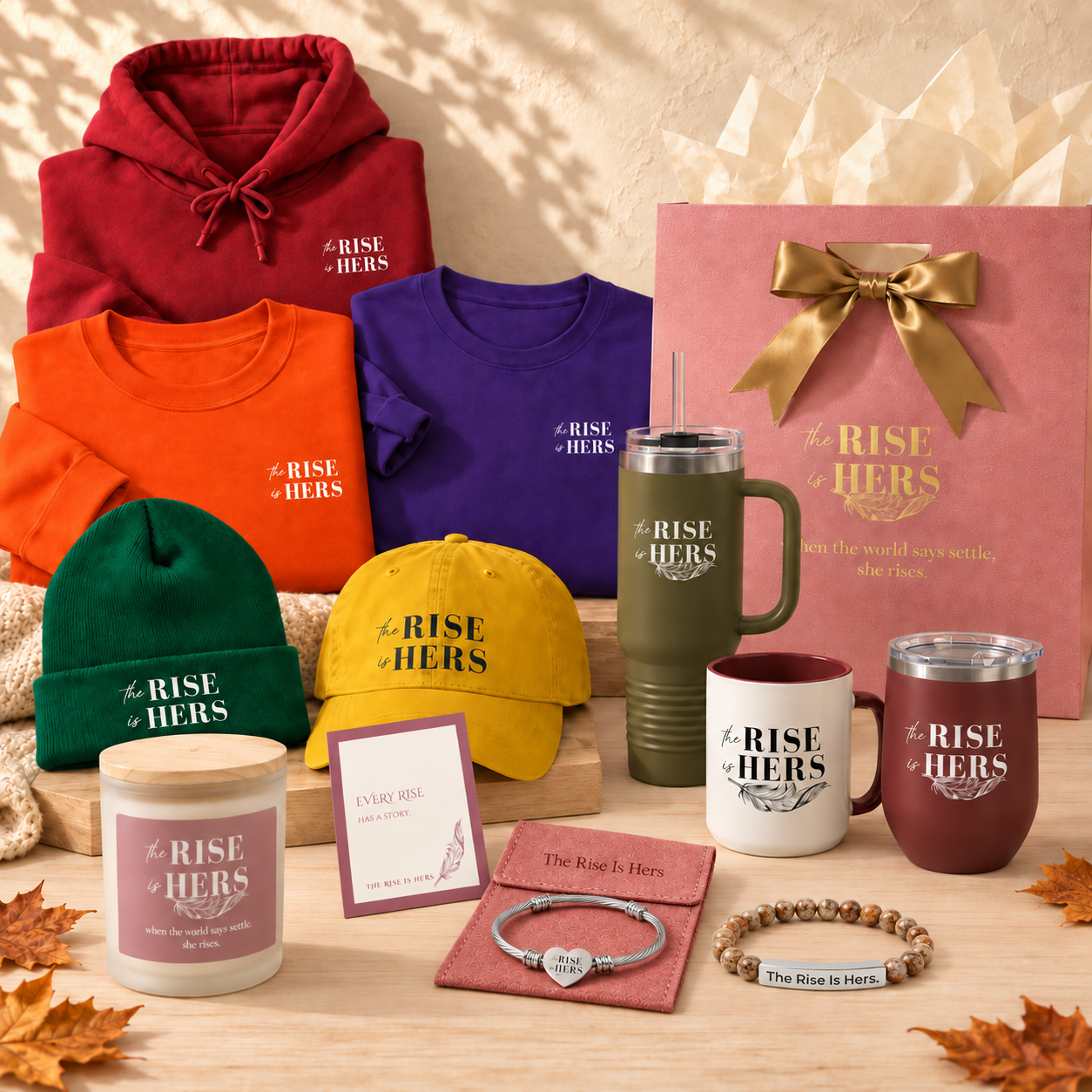 The Rise Is Hers fall gift collection with embroidered apparel, drinkware, candles, bracelets, and a pink gift bag with gold ribbon.