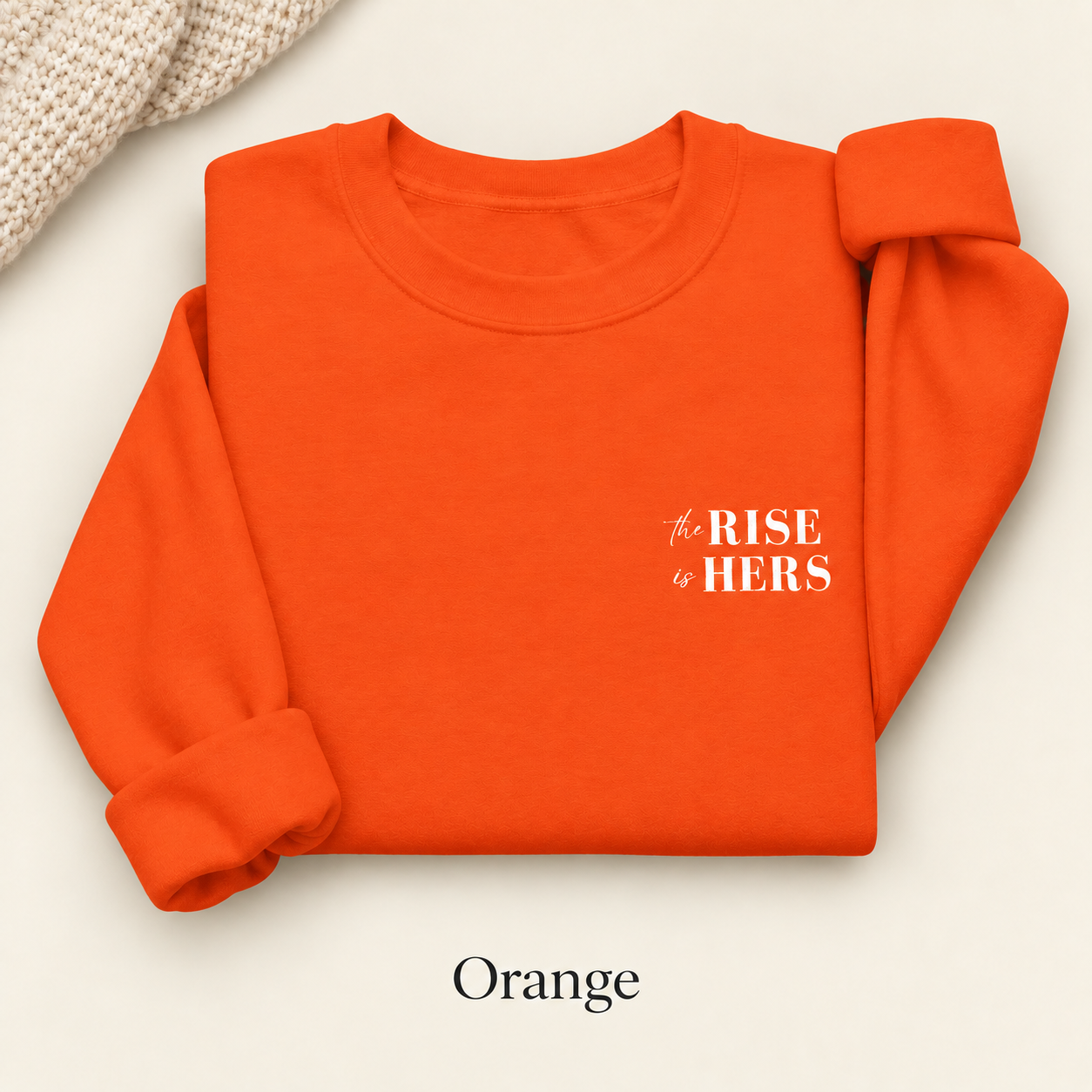 Orange The Rise Is Hers crewneck sweatshirt with an embroidered left-chest logo, folded on an ivory background.