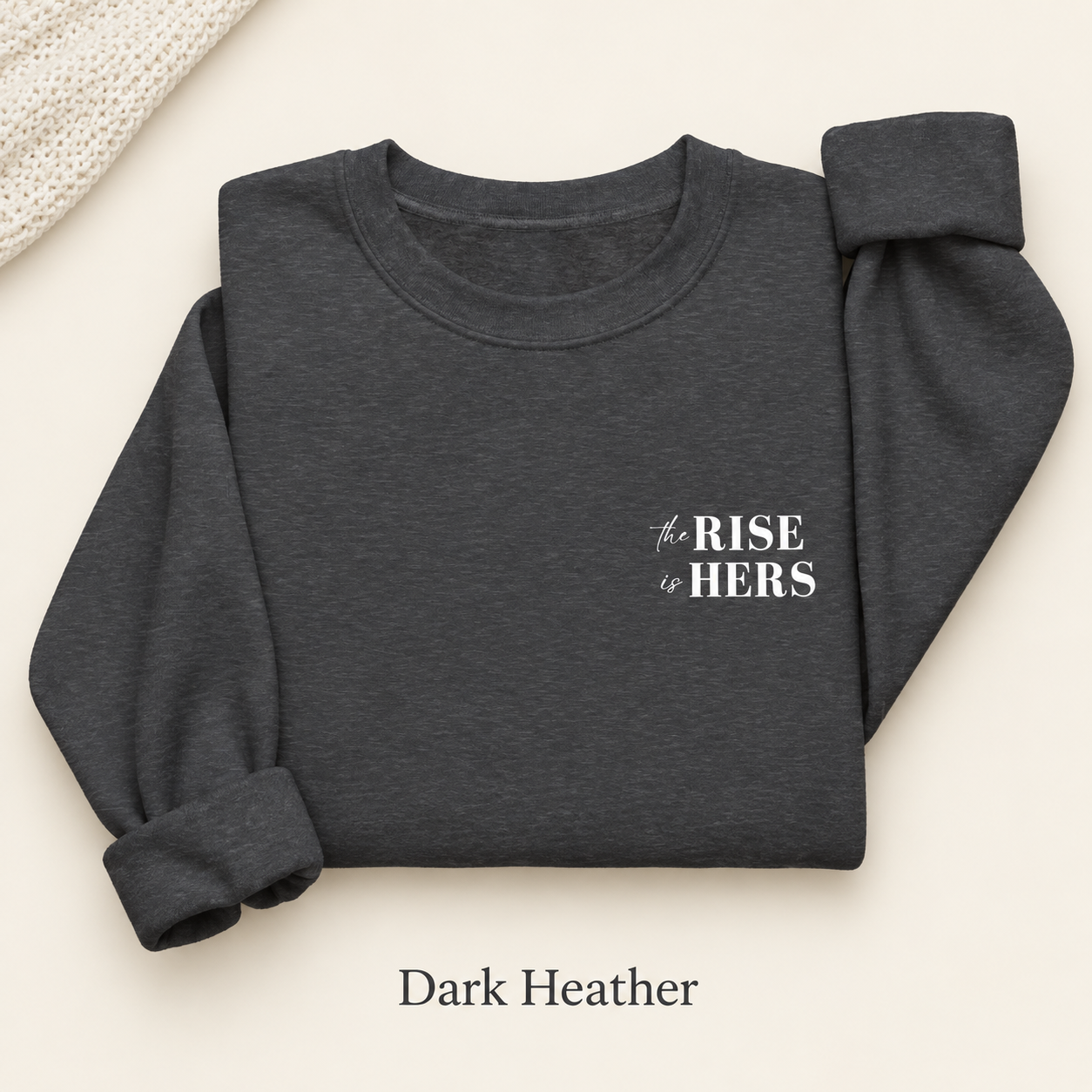 Dark Heather gray The Rise Is Hers crewneck sweatshirt with an embroidered left-chest logo, folded on an ivory background.