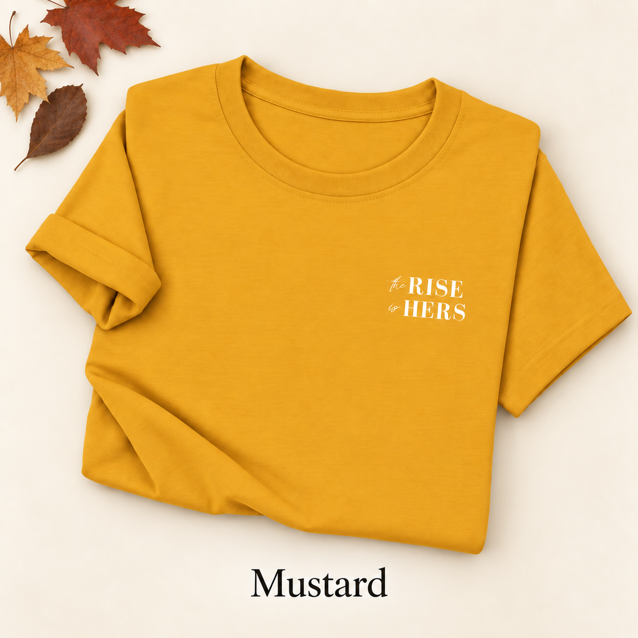 Mustard yellow The Rise Is Hers T-shirt with an embroidered left-chest logo, folded on an ivory background with autumn leaves.
