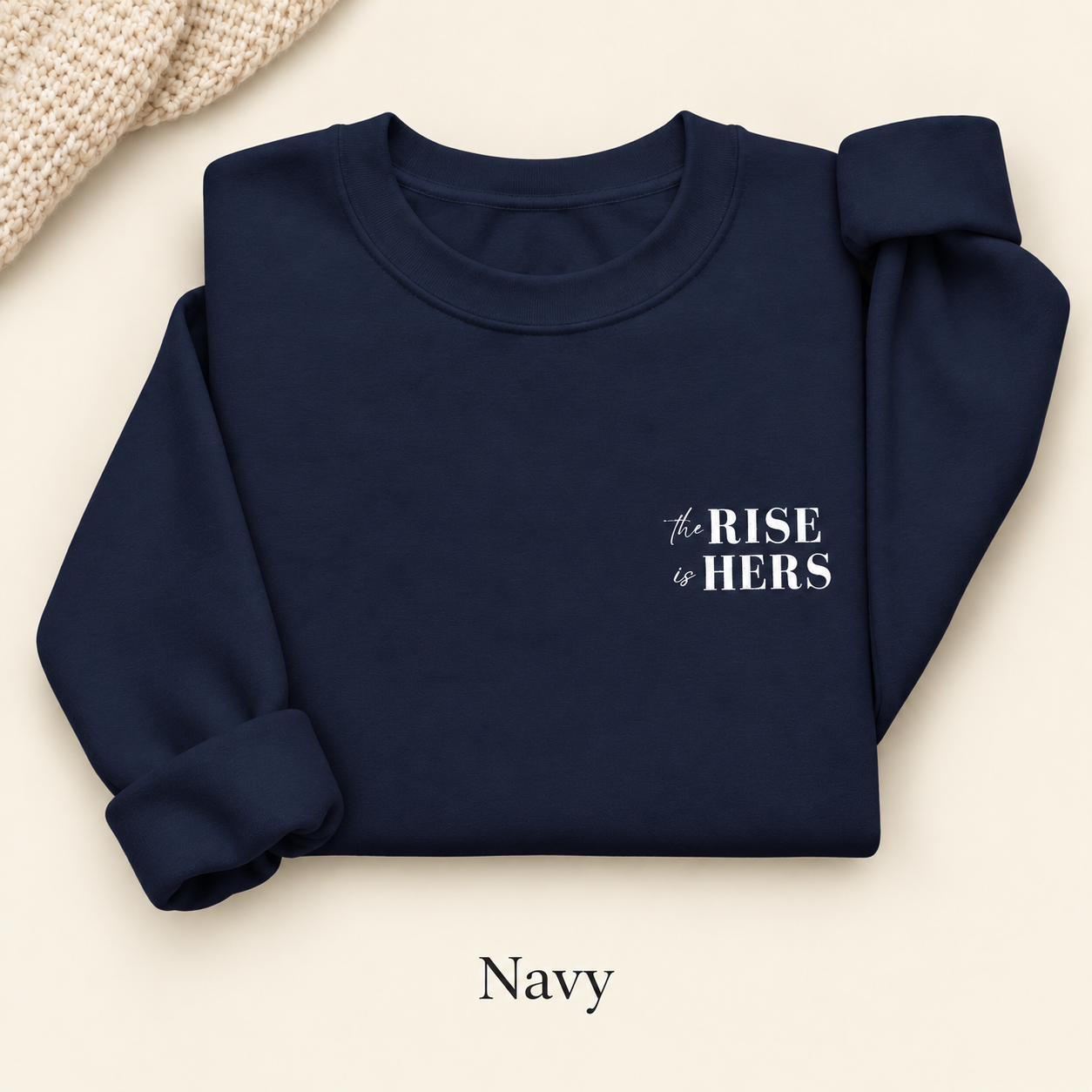 Navy blue The Rise Is Hers crewneck sweatshirt with an embroidered left-chest logo, folded on an ivory background.