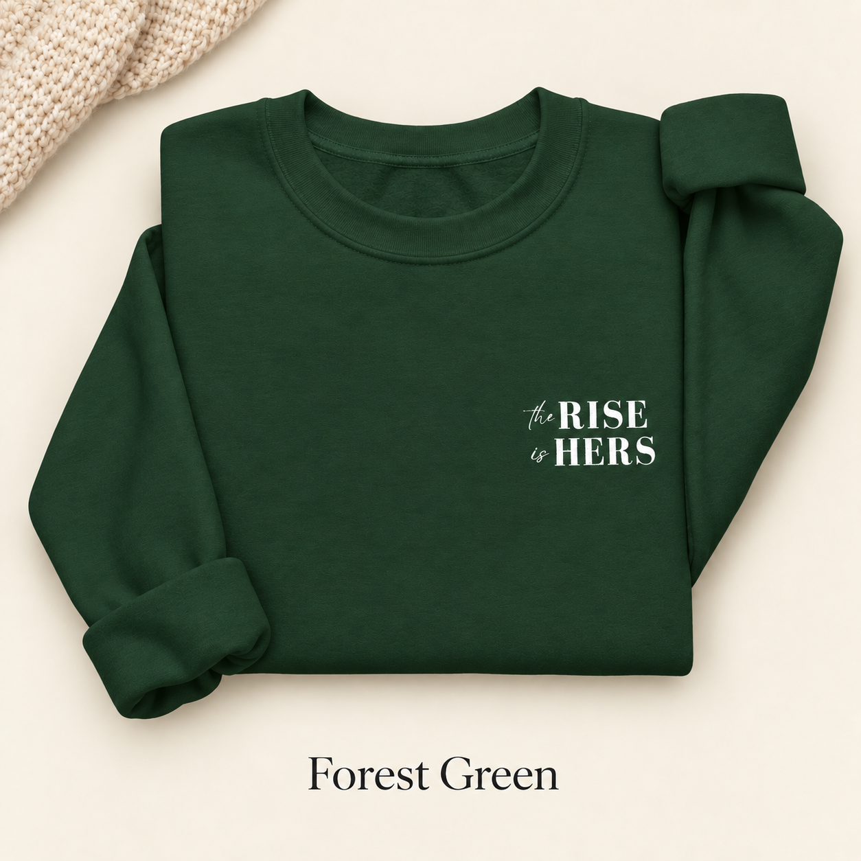 Forest Green The Rise Is Hers crewneck sweatshirt with an embroidered left-chest logo, folded on an ivory background.