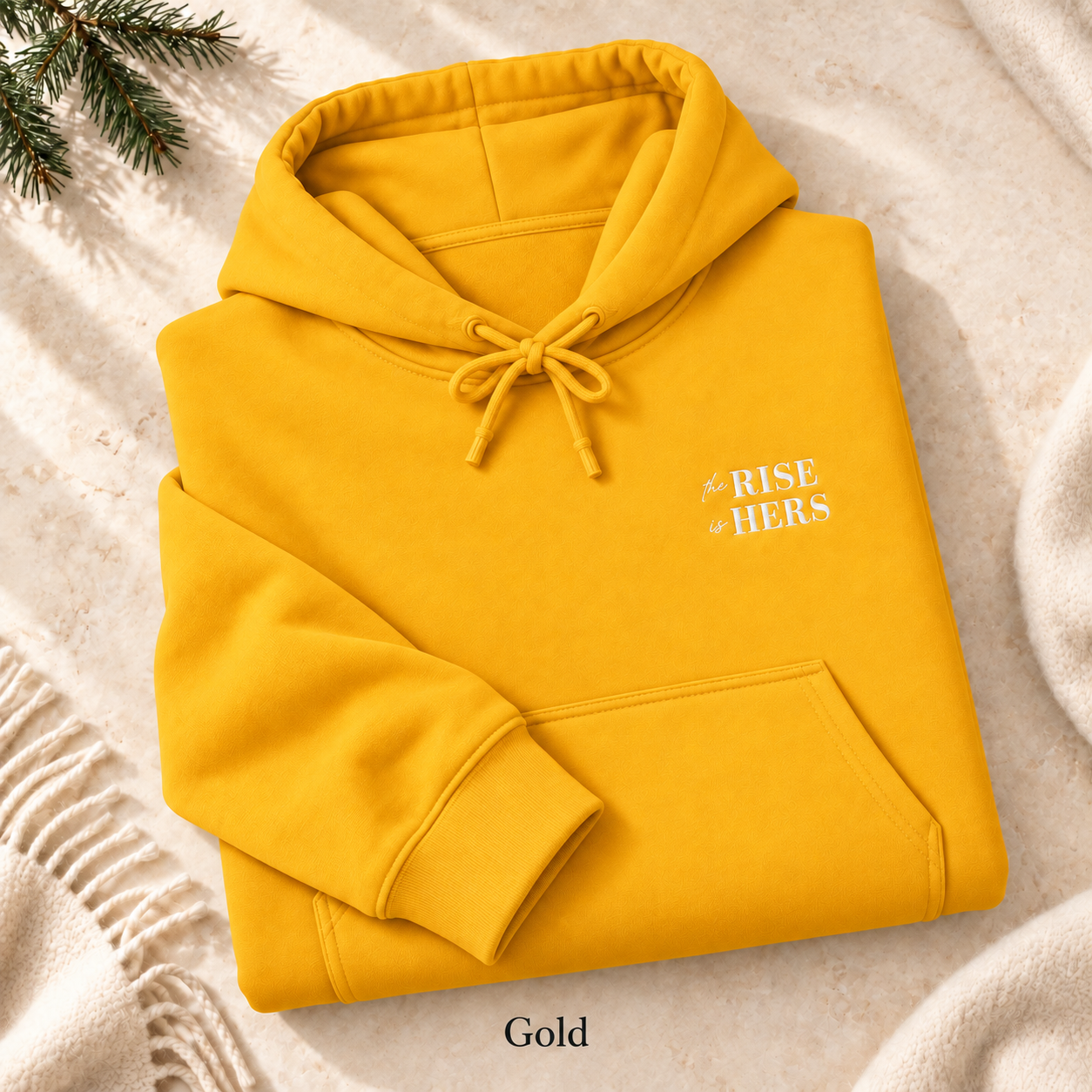 Gold The Rise Is Hers hoodie with an embroidered left-chest logo, folded on cream fabric.