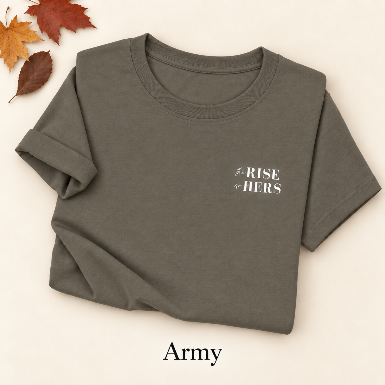 Army green The Rise Is Hers T-shirt with an embroidered left-chest logo, folded on an ivory background with autumn leaves.