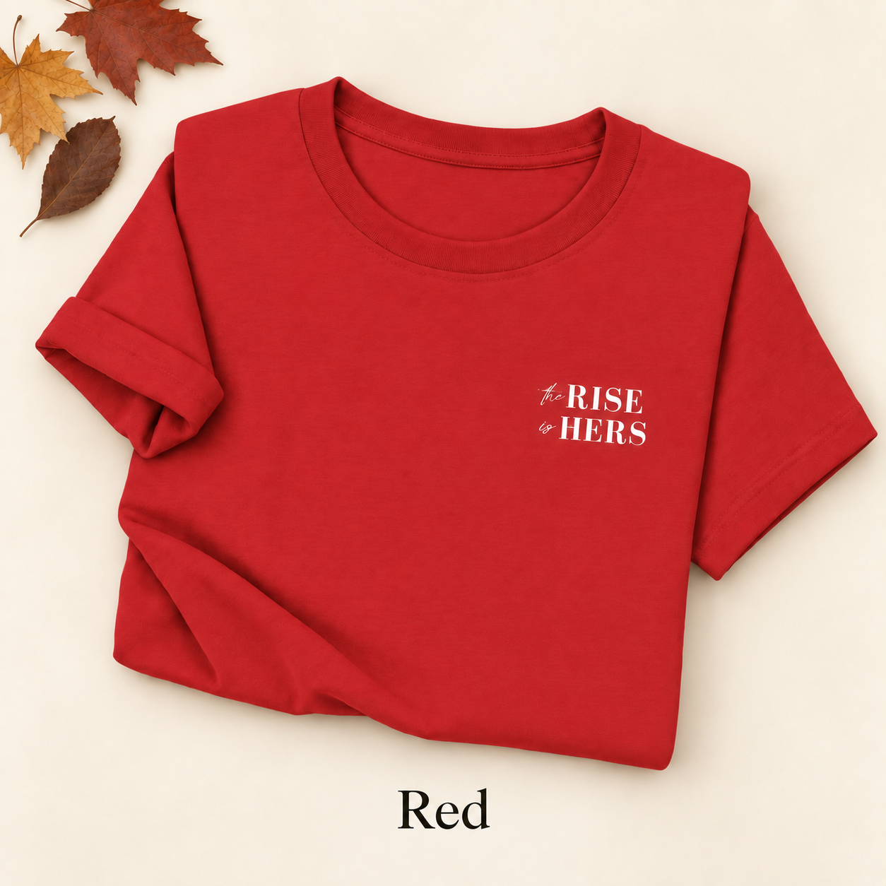 Red The Rise Is Hers T-shirt with an embroidered left-chest logo, folded on an ivory background with autumn leaves.