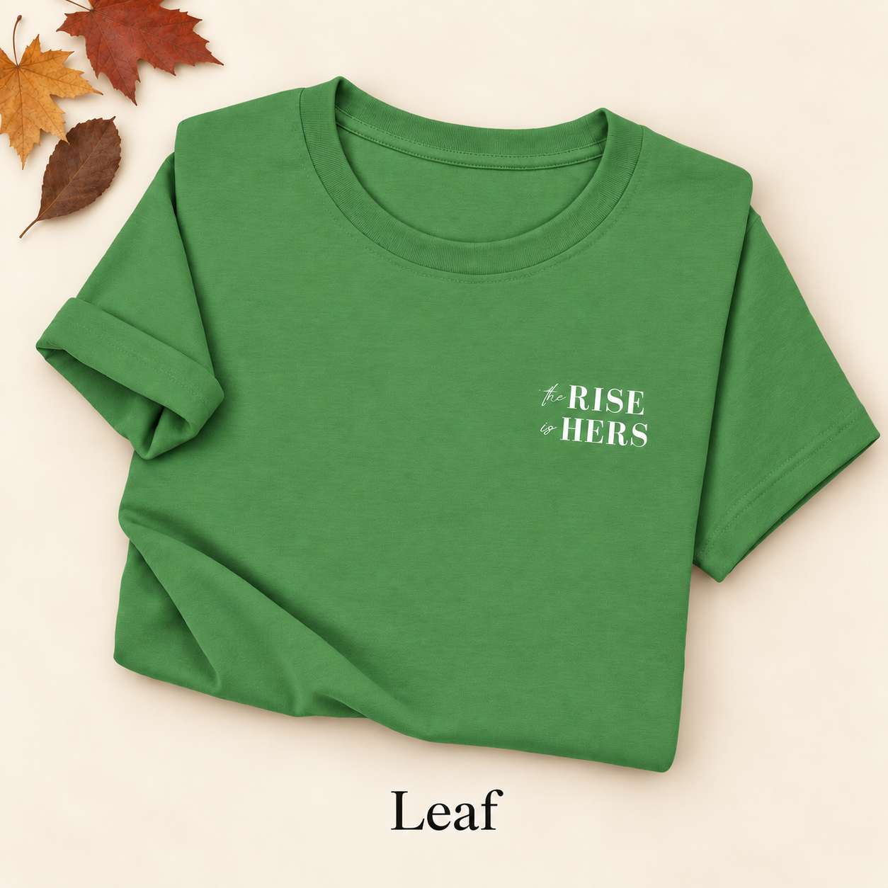 Leaf green The Rise Is Hers T-shirt with an embroidered left-chest logo, folded on an ivory background with autumn leaves.