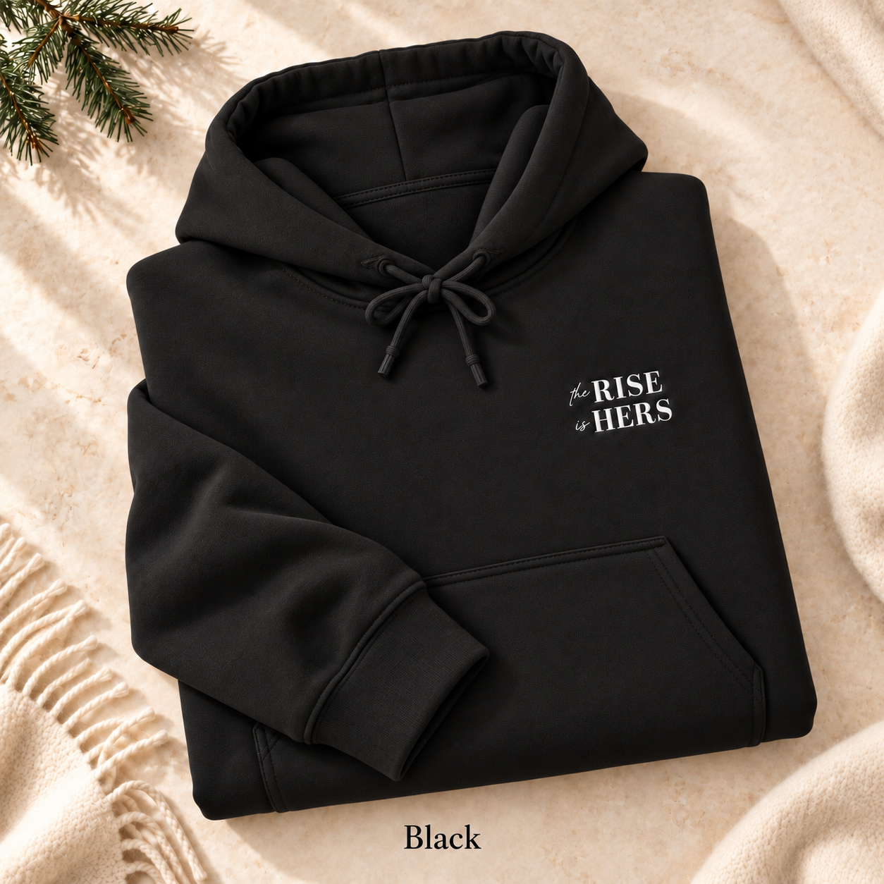 Black The Rise Is Hers hoodie with an embroidered left-chest logo, folded on cream fabric.