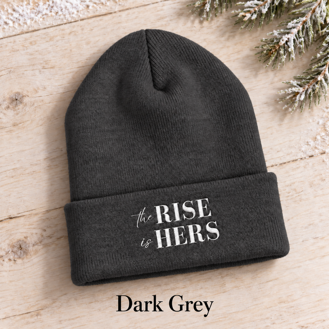 Dark Grey The Rise Is Hers cuffed knit beanie with an embroidered logo, displayed on a light background beside an evergreen branch.
