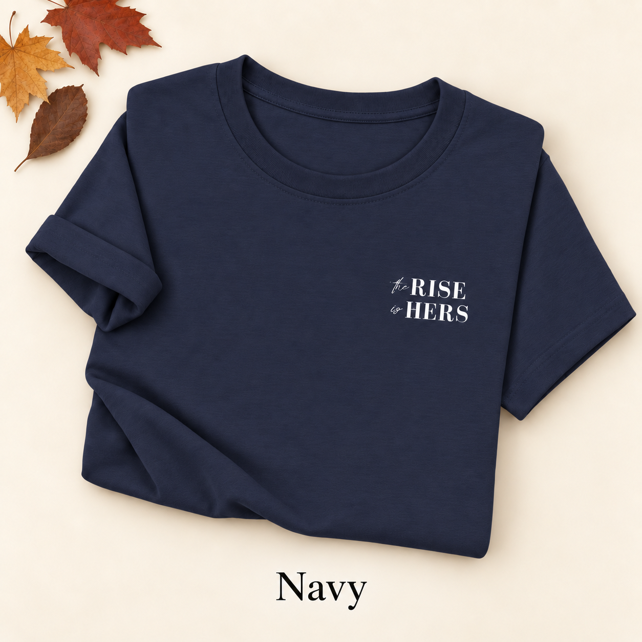 Navy blue The Rise Is Hers T-shirt with an embroidered left-chest logo, folded on an ivory background with autumn leaves.
