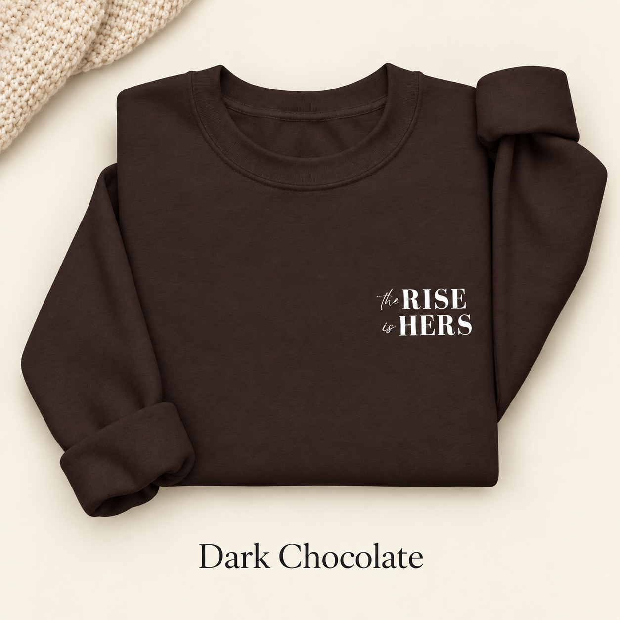 Dark Chocolate brown The Rise Is Hers crewneck sweatshirt with an embroidered left-chest logo, folded on an ivory background.