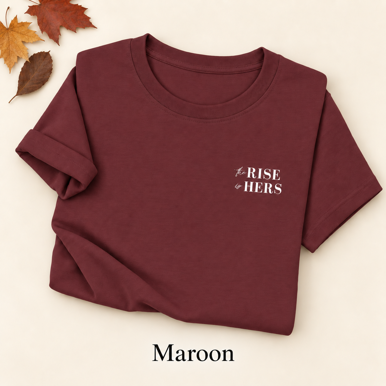 Maroon The Rise Is Hers T-shirt with an embroidered left-chest logo, folded on an ivory background with autumn leaves.