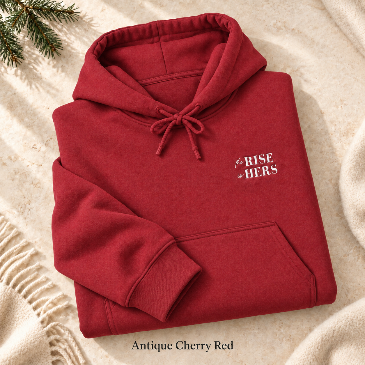 Antique Cherry Red The Rise Is Hers hoodie with an embroidered left-chest logo, folded on cream fabric.