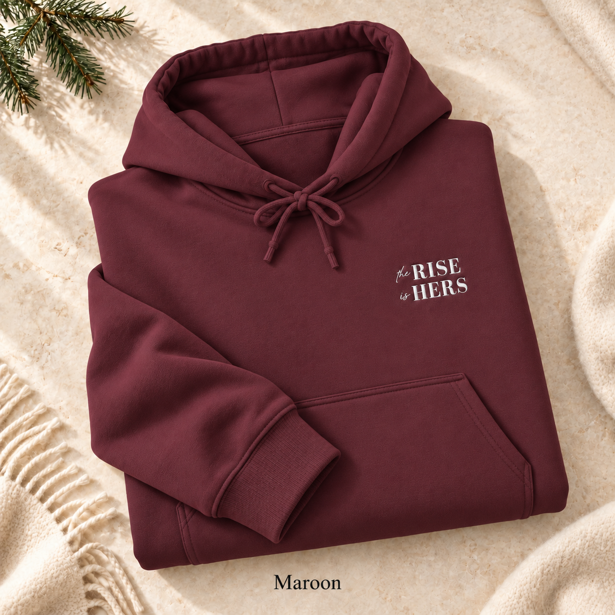 Maroon The Rise Is Hers hoodie with an embroidered left-chest logo, folded on cream fabric.