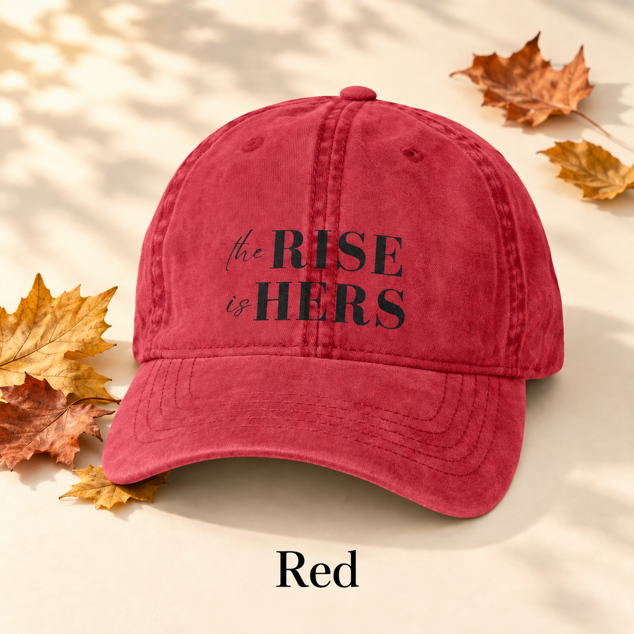 Red The Rise Is Hers baseball hat with an embroidered front logo, displayed beside autumn leaves.