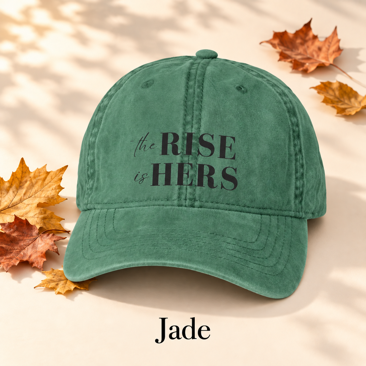 Jade green The Rise Is Hers baseball hat with an embroidered front logo, displayed beside autumn leaves.